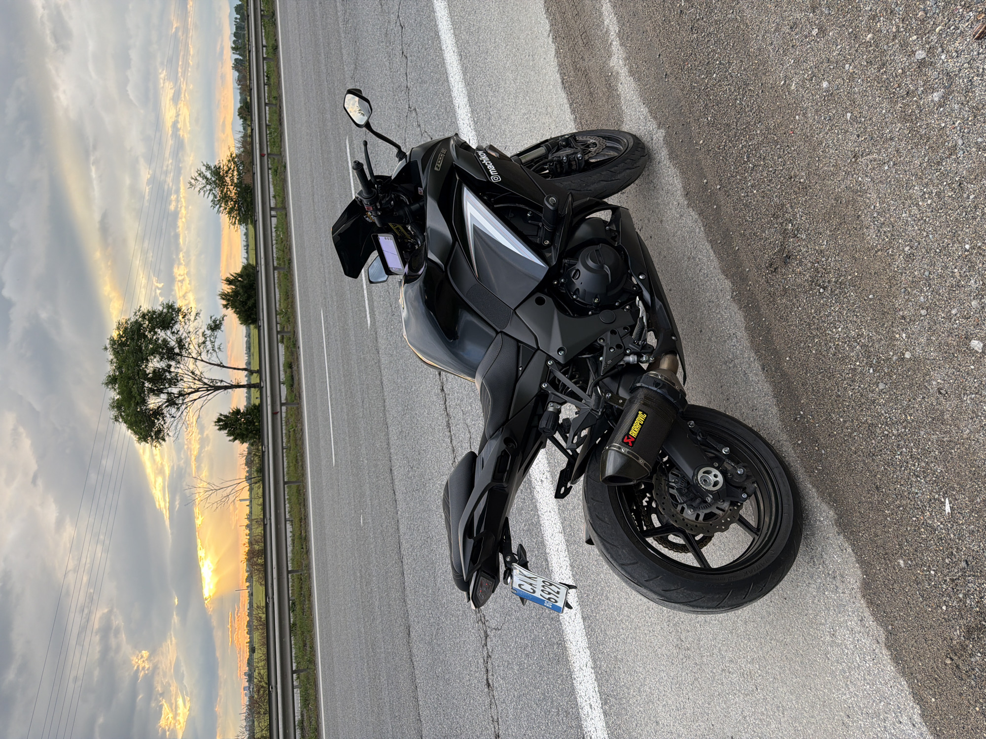 The image shows a black Kawasaki Z1000 motorcycle parked on the shoulder of a road near Kazichene, Bulgaria. The sun is setting, creating a dramatic sky. The motorcycle is facing towards the right, and the rear license plate is visible. The location is identifiable by the landscape and roadside features typical of the area.