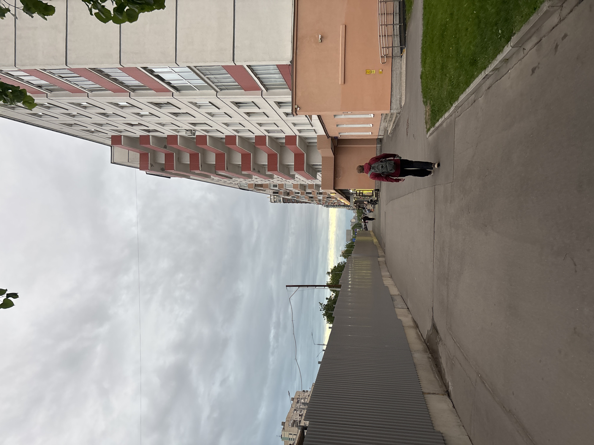 A street scene in Unknown, Unknown. A person with a backpack walks along a sidewalk next to a tall building with red balconies. A corrugated metal fence runs along the other side of the sidewalk. The sky is cloudy.
