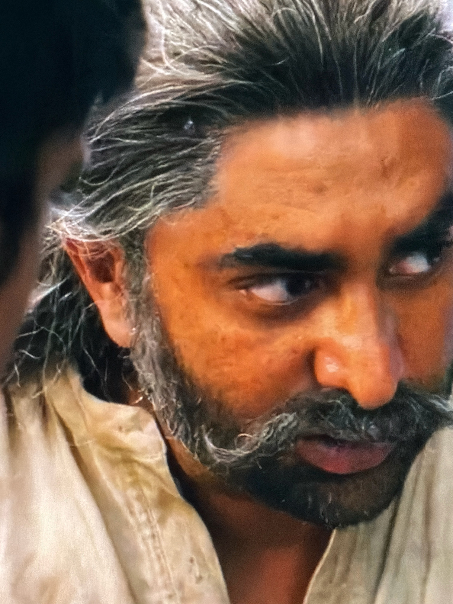 The image is a close-up, slightly blurry photograph of a man's face. He has dark skin, long greyish-black hair that is pulled back, a prominent mustache, and a beard. His eyes are looking off to the side, and his mouth is slightly open. He is wearing a light-colored garment, possibly a shirt or robe. The background is dark and out of focus. There is no visible indication of the location being Agachaul, Russia. The image focuses solely on the man's facial features.