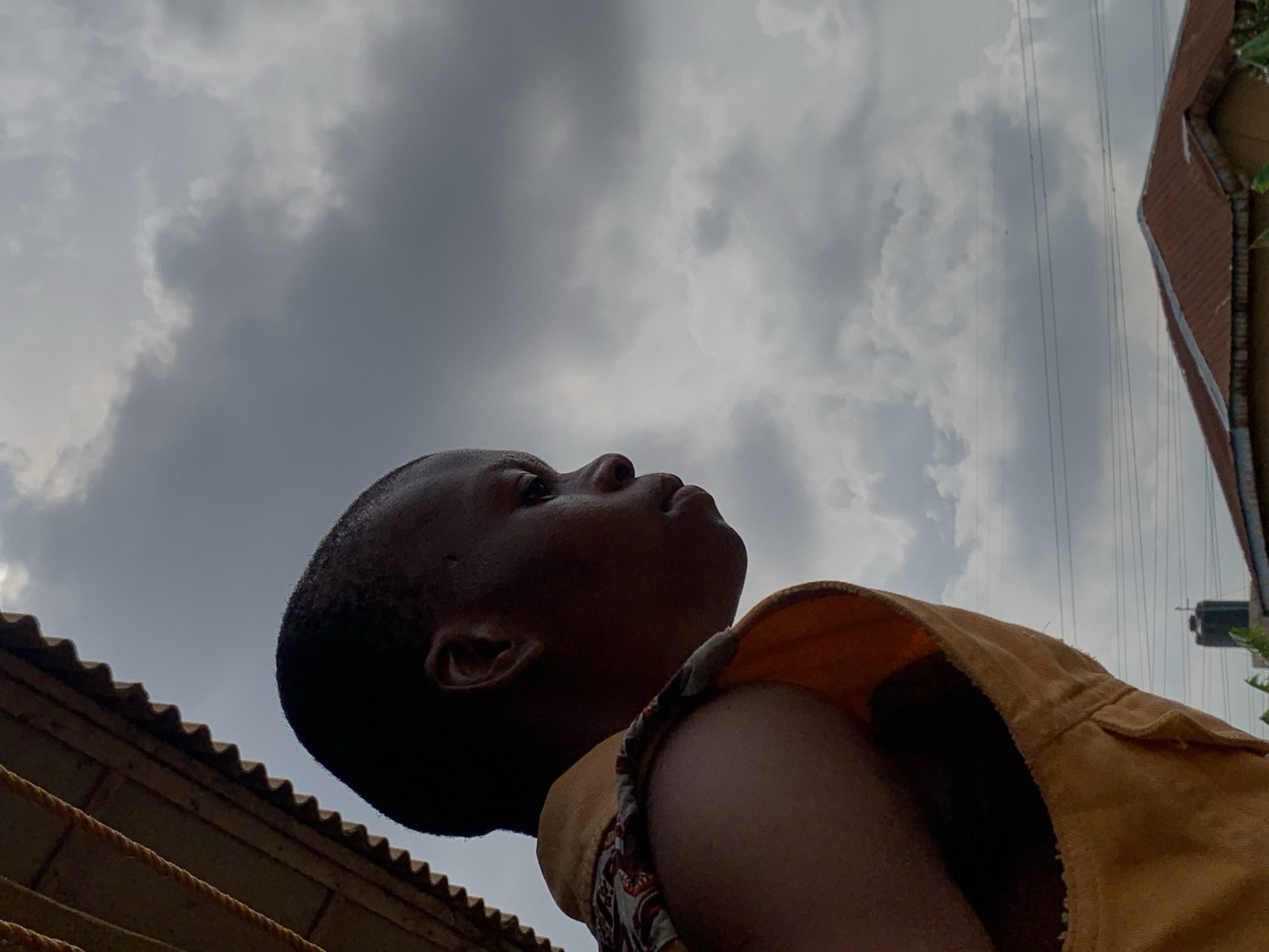 A low-angle shot features a person with dark skin, looking upward, possibly into a cloudy sky. The person is wearing a tan vest over a patterned shirt. The background includes power lines, the edge of a building with a corrugated roof, and a glimpse of another building in the distance. Although the image is not explicitly geo-tagged or described, if the location is indeed Ikorodu, Nigeria as specified, the scene suggests an urban environment in the tropics, characterized by a mix of residential and commercial structures and overhead power infrastructure.
