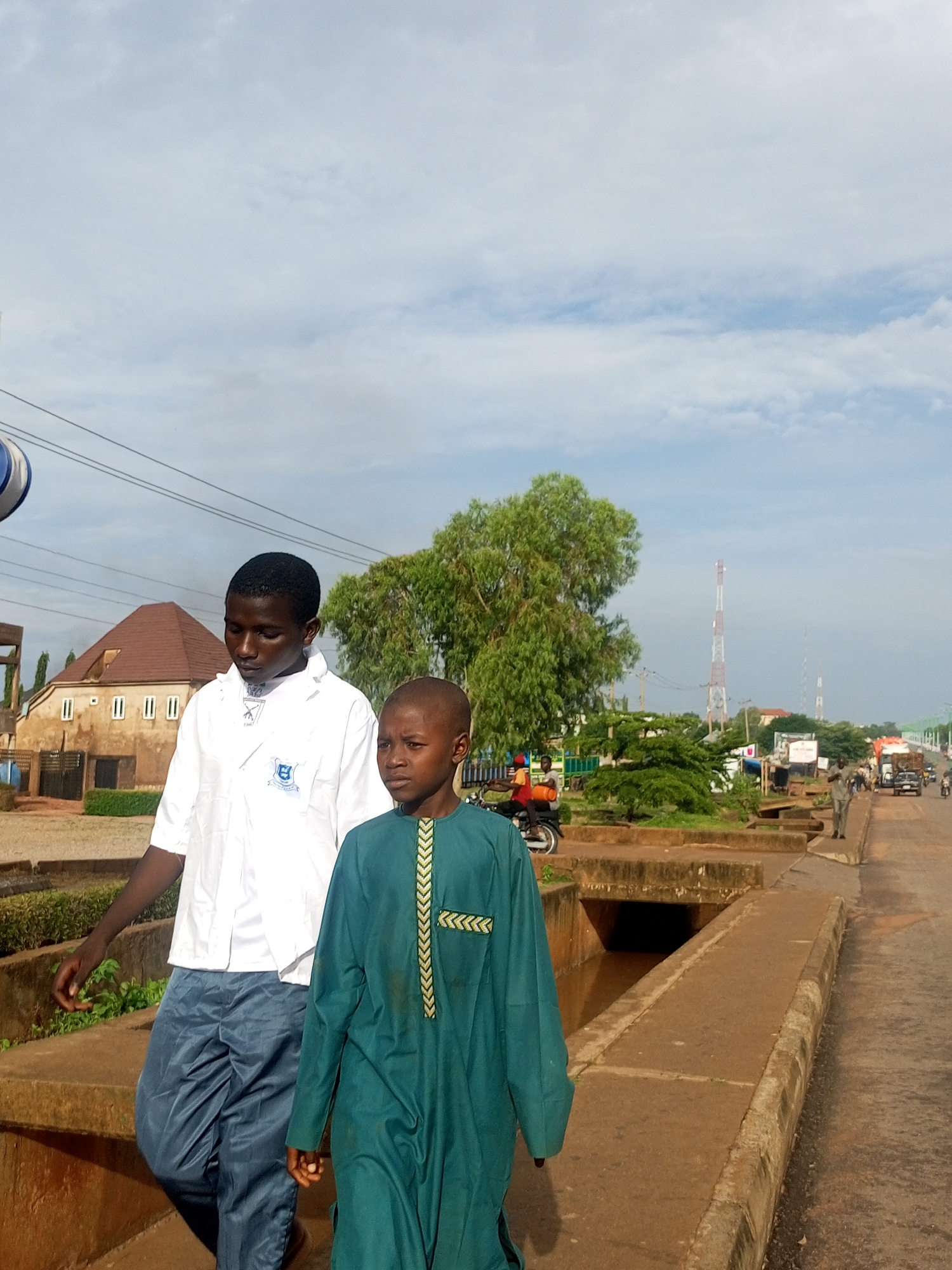 Two young males are walking on a sidewalk next to a road. The male on the left is wearing a white button-down shirt with a blue logo and blue pants. The male on the right is wearing a green robe with gold trim. They are in an urban environment with buildings, trees, and a drainage ditch. In the background, there are cell towers, vehicles, and other pedestrians. The sky is cloudy.