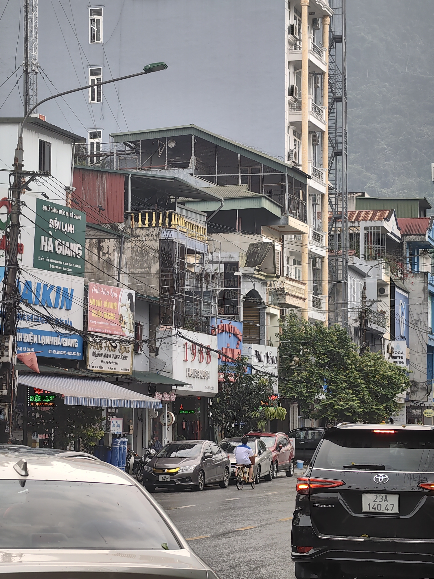 The image captures a street scene in Thành phố Hà Giang, Vietnam, on what appears to be a cloudy day. In the foreground, parts of two cars are visible: the rear of a black Toyota Fortuner with the license plate "23A 140.47" is positioned on the right, and a portion of a silver car is on the left. A man on a bicycle is riding in the lane between the two cars.
The background is filled with a dense urban environment, characterized by closely packed buildings of varying heights, styles, and colors. The buildings feature multiple signs and advertisements, some of which are partially obscured by power lines. The signs display Vietnamese script and include the names "HA GIANG", "DAIKIN", "Ánh Hòa Spa", and "1988 HAIRSALON".
Higher in the frame, a taller, pale grey building dominates the view, flanked by a vertical metal staircase on one side. In the distance, the blurred silhouette of a mountain range is visible through the overcast sky.