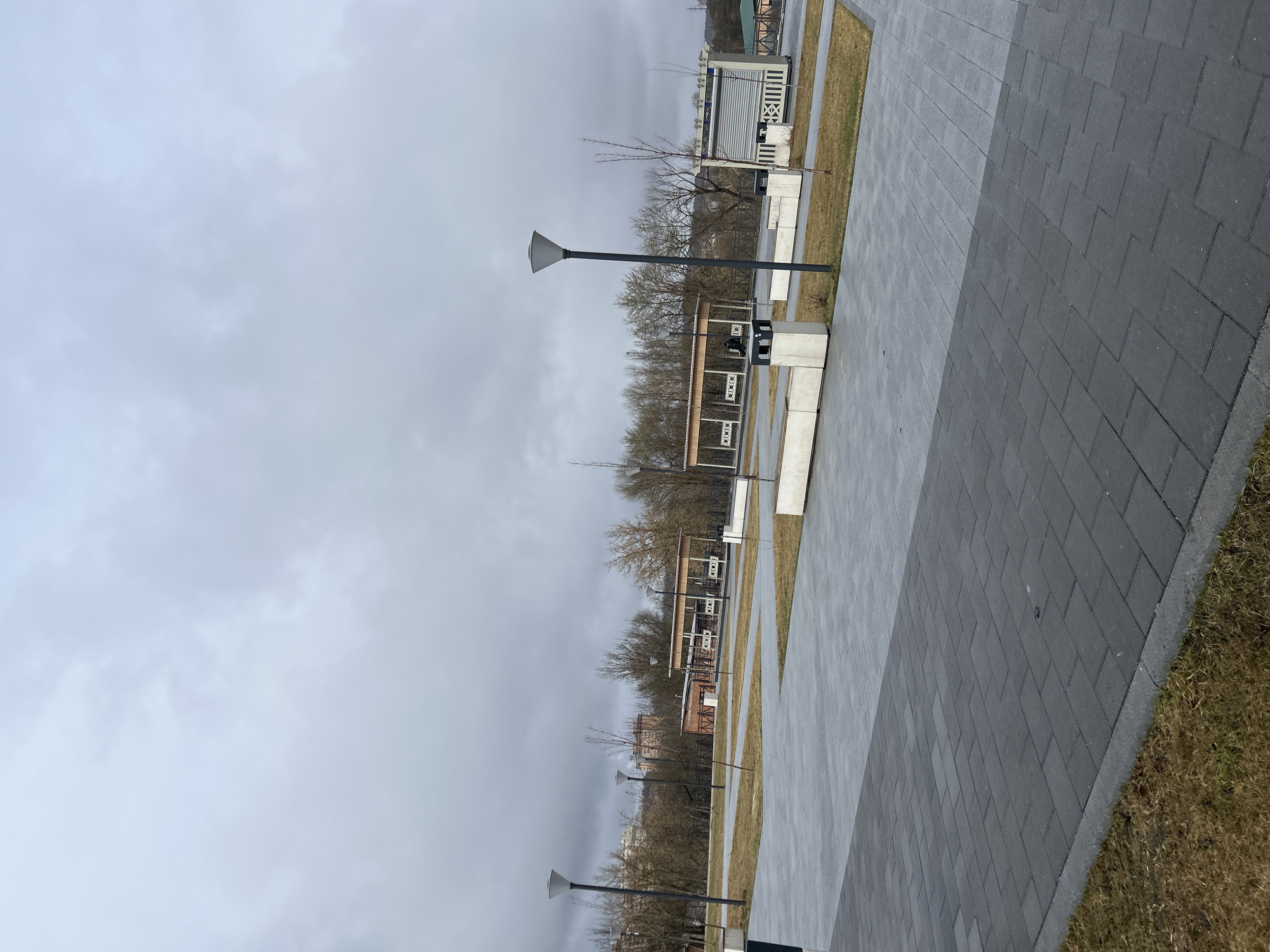 The image depicts a paved walkway in Klin, Russia, under a cloudy sky. The walkway is constructed of gray rectangular pavers, with two distinct shades. To the right of the walkway is a strip of dry grass.
Parallel to the walkway, and separated by a strip of grass, are a series of small structures. Each structure consists of a base with concrete walls, and a decorative structure above with a white geometric pattern. There are benches and trash cans next to the structures.
Bare trees line the area behind the structures. Further in the background are apartment buildings.
Streetlights with conical lamps are spaced along the walkway.