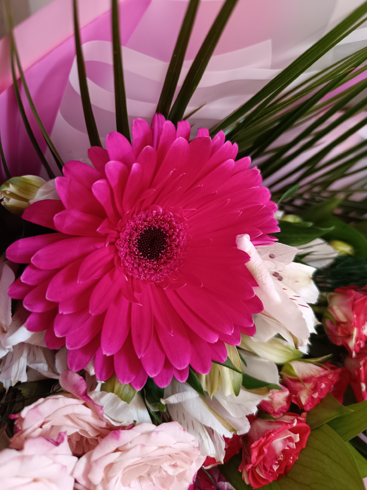 The image depicts a close-up of a flower bouquet. Prominently featured is a vibrant pink gerbera daisy with a dark brown center. Surrounding the gerbera are white alstroemeria lilies and roses, some with pink and red hues. Green, linear leaves extend upwards behind the flowers, and pink and white decorative wrapping paper is also visible in the background.