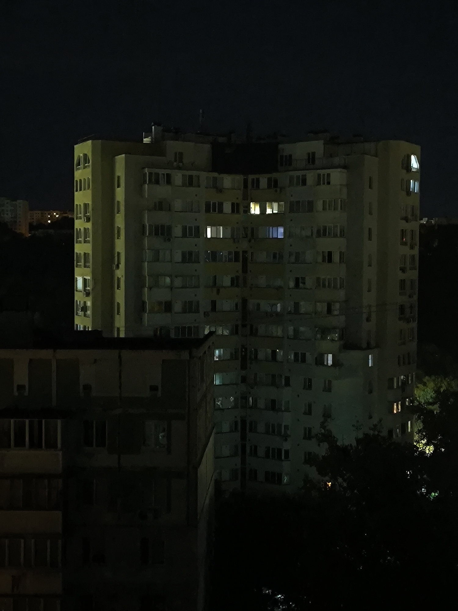 The image depicts a nighttime view of a large, multi-story residential building in Chișinău, Moldova. The building, painted in a light yellow or cream color, is illuminated by the lights from many of its windows, suggesting occupancy. It has a distinctive rounded corner on the left side. The structure is quite tall, with numerous rows of windows and balconies. In the foreground, a darker, lower building is partially visible, its windows largely dark. Trees and foliage are present in the lower right and on the left, partially obscuring the view of the buildings. The sky above is dark, indicative of nighttime. The overall impression is an urban landscape at night, focusing on residential architecture.
