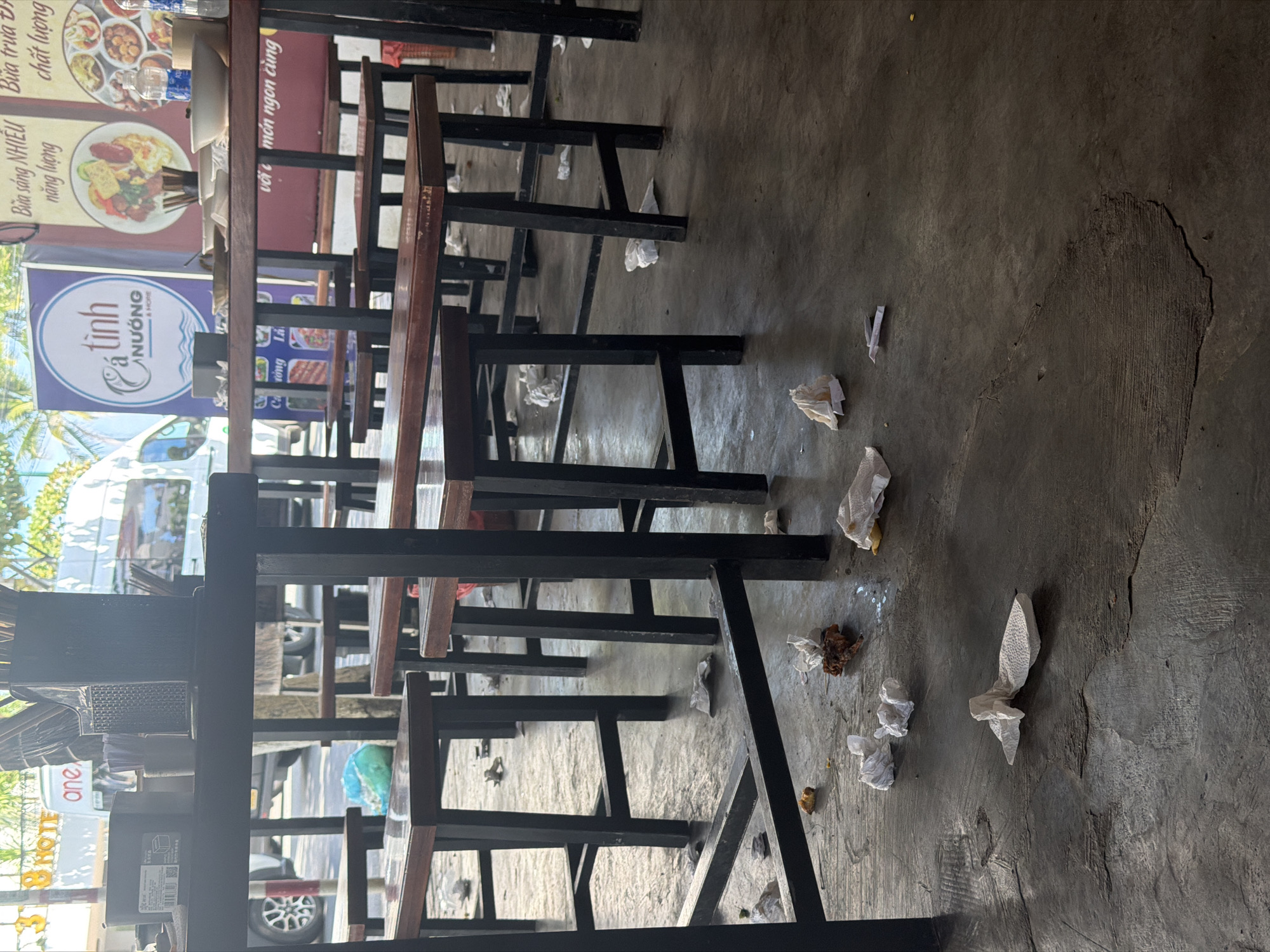 The image captures a ground-level view inside a casual restaurant or eatery in Nha Trang Ward, Vietnam, likely after a busy period. The concrete floor, occupying most of the frame, appears worn, with visible cracks and patches, and is significantly littered with crumpled white paper napkins, small food scraps, and a blue plastic bag, indicating a lack of immediate cleaning.
Numerous tables and chairs, characterized by dark metal frames and wooden tops and seats, are arranged in rows, suggesting a functional, unpretentious dining setup. No people are visible, but the scattered debris strongly implies recent activity.
Through an open front or large window, bright daylight streams in, revealing a street scene with vehicles, including a white van, and green trees/foliage. A building with the partial text "one" and "HOTEL" is visible in the background reflection or through the opening.
Several banners displayed in the establishment feature Vietnamese text. A prominent blue sign with a fish logo reads "Cá tinh NƯỚNG & HOME". Other banners advertise meals, with phrases like "Bữa sáng NHIỀU năng lượng" (Breakfast with lots of energy), "Bữa trưa chất lượng" (Quality lunch), and "với món ngon cùng" (with delicious dishes with). A plastic water bottle rests on one of the tables. The overall atmosphere suggests a well-used local dining spot with a casual, rather messy, environment.