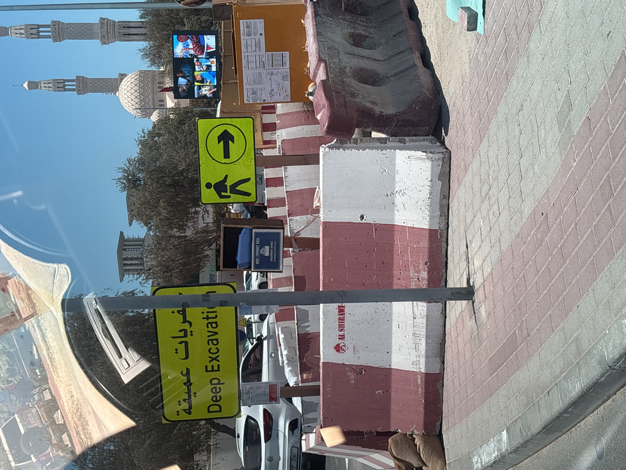 The image depicts a street scene in Dubai, United Arab Emirates, with a focus on a construction site. In the foreground, there are concrete barriers painted with red and white stripes, commonly used for traffic control and safety. One of these barriers features the logo "AL SHIRAWI". A dark sandbag is visible to the left of the barriers.
A prominent yellow sign with Arabic text and the words "Deep Excavation" in English is mounted on a metal pole. Next to it, a green sign with a pedestrian silhouette and an arrow pointing to the right indicates a designated walkway.
In the background, several vehicles are parked, including a white SUV with a license plate visible. Beyond the construction site, the iconic architecture of Dubai can be seen, including a mosque with a dome and minarets, suggesting a location within the city. A large LED screen displaying multiple images of people is also visible. The environment is bathed in sunlight, indicating a clear day. The ground is paved with interlocking bricks.