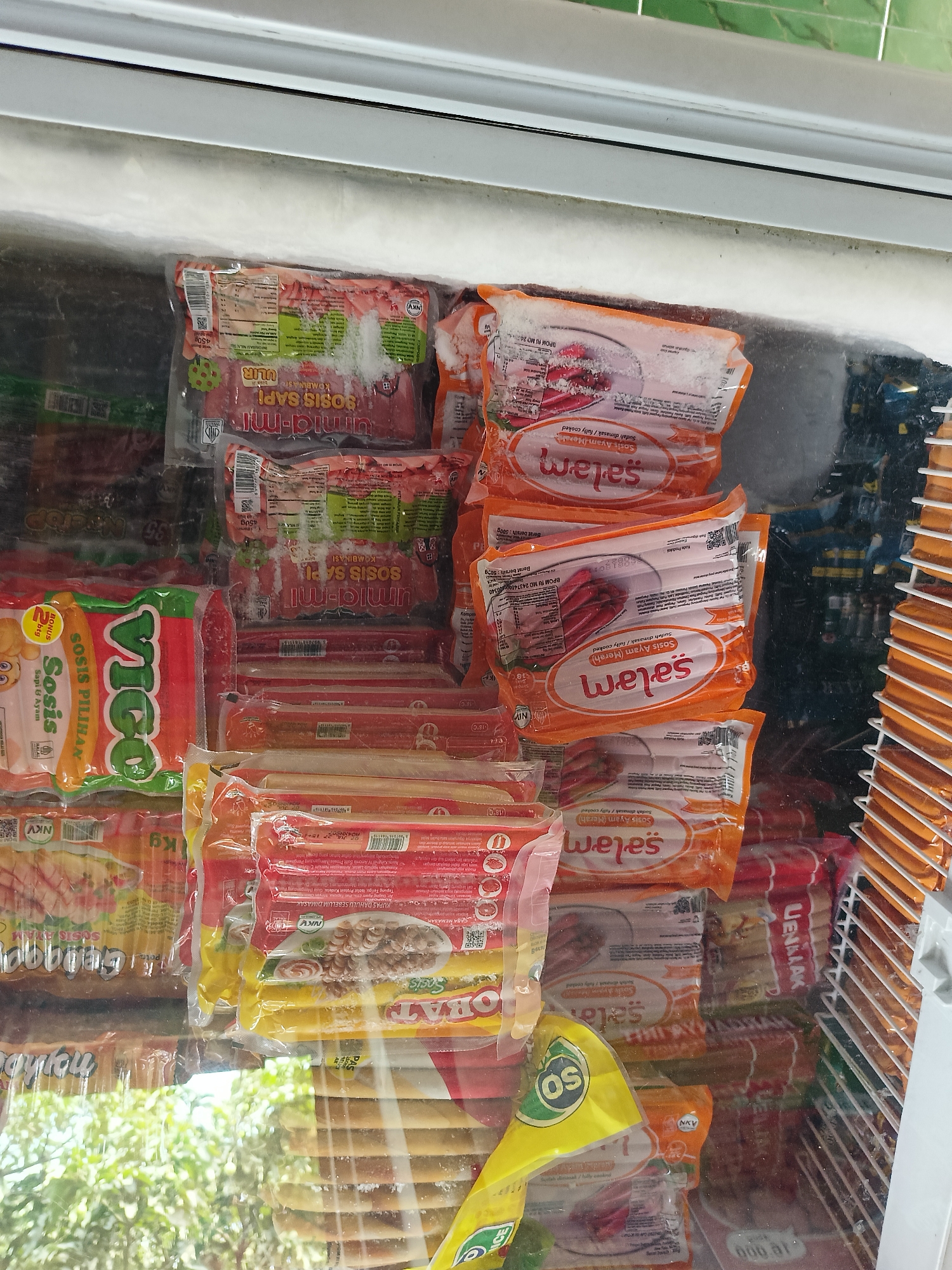 The image shows a freezer chest filled with various packaged sausages and processed meat products, primarily in a retail setting. The products are predominantly in plastic packaging with prominent branding and nutritional information, suggesting they are for sale. Some of the packaging is covered in frost, indicating the freezer's function. The items include different types of sausages, such as "Sosis Sapi" (beef sausage) and "Sosis Ayam" (chicken sausage), with brands like "Umami-mi" and "Vigo" visible. There are also items labeled "Salam" which appears to be a type of sausage or processed meat. In the background, more packaged food items are visible, along with metal shelving units. The overall scene is that of a food storage or retail display within a commercial establishment. The image is taken from a slightly elevated angle looking down into the freezer. There are no people visible in the image. The environment appears to be indoors, with reflections on the freezer glass suggesting an artificial light source and possibly other merchandise or walls in the vicinity.