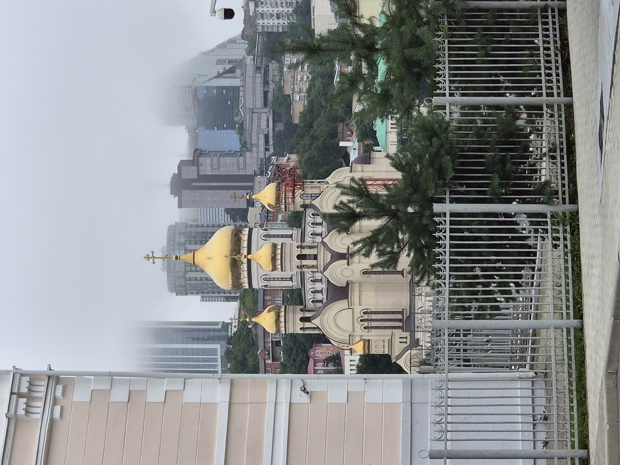 The image shows a view of the city of Vladivostok, Russia, with a prominent Orthodox church featuring golden domes in the foreground. The church is a large, ornate structure with multiple onion domes, the largest of which is topped with a golden cross. Behind the church, the city skyline is visible, characterized by modern high-rise buildings, some of which are partially obscured by fog or mist. The buildings are a mix of glass and concrete structures. The landscape is hilly, with trees and smaller residential buildings dotting the slopes between the church and the modern city. In the immediate foreground, a decorative metal fence runs horizontally across the image, with a paved walkway and a curb visible at the very bottom. Green pine trees are interspersed in front of the fence and partially obscure the view of the church. The overall atmosphere is overcast, with the sky appearing a uniform grey. A security camera is visible mounted on a pole on the right side of the image. The building on the far left is a light-colored structure with architectural details at the top, and it partially frames the scene.