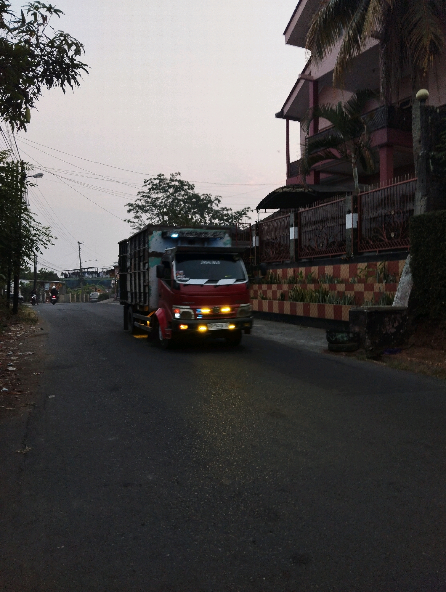 A medium shot captures a red dump truck with a green cargo container driving on a paved road in what appears to be a residential area during twilight. The truck is in motion, indicated by motion blur, and its headlights are illuminated. The road is asphalt and shows some wear and tear, with patches of dirt and debris along the side.
To the right of the road, a two-story building with a pink facade and dark railings is visible. In front of the building, there is a decorative fence with ornate metalwork and a tiled wall featuring a checkered pattern of red and beige tiles. Green plants are planted in sections along the tiled wall. A tall palm tree grows near the building.
To the left of the road, there are trees with dark green foliage and electrical wires strung across poles. Further down the road, two motorcycles are also visible, suggesting the presence of other vehicles. The sky is overcast and a pale gray, indicating low light conditions. The overall scene suggests a street in Salatiga, Indonesia, characterized by a mix of residential buildings, infrastructure, and vegetation.