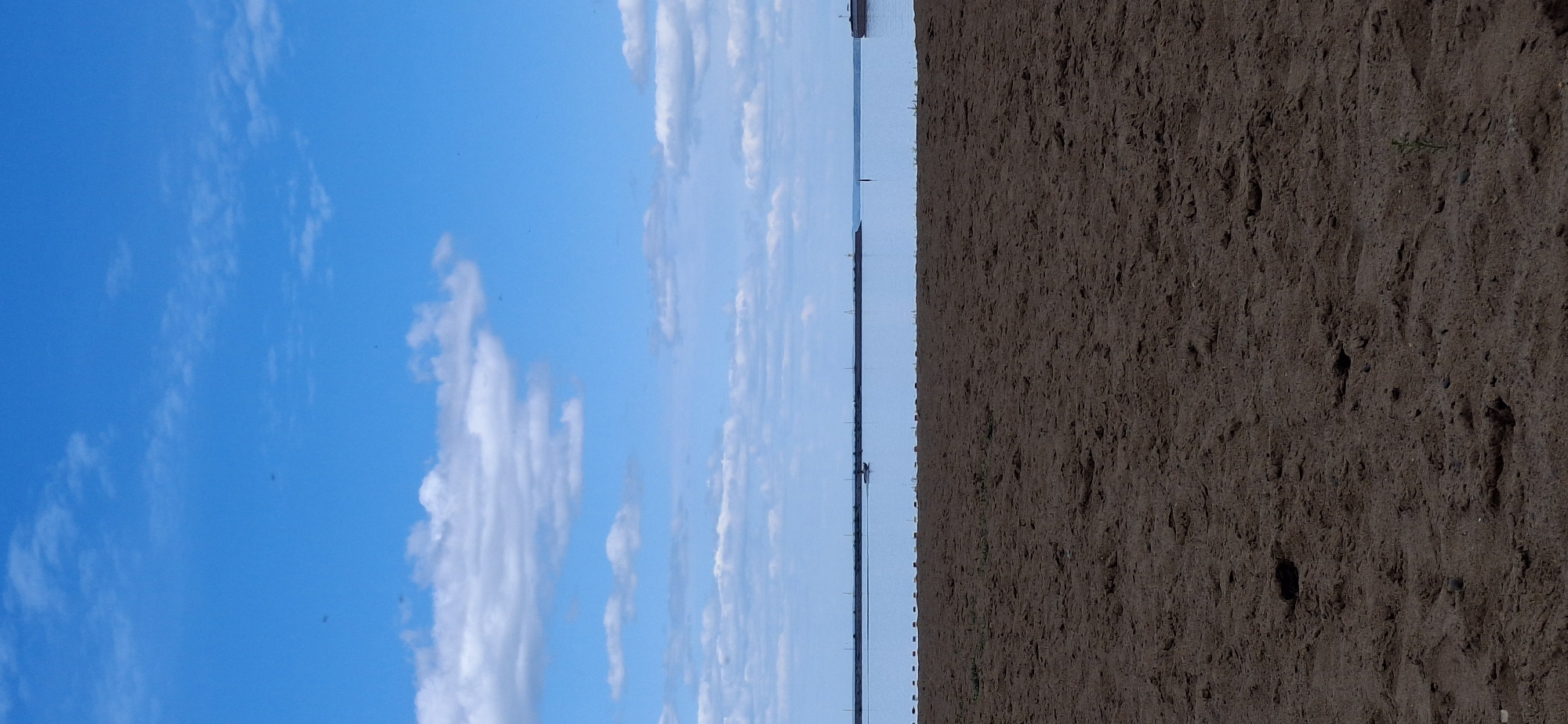 This image depicts a coastal or lakeside scene in Chaikovsky, Russia, characterized by a sandy foreground, a body of water in the midground, and a blue sky with clouds in the background.
The foreground occupies approximately the lower half of the image and consists of dark brown, granular sand, exhibiting minor textural variations and small indentations. A single slender green plant is visible near the bottom right edge.
A calm expanse of light blue water occupies the central horizontal band of the image. A series of evenly spaced, dark, rectangular markers or buoys delineate the immediate edge of the water from the sand. Along the distant horizon, a long, dark, linear structure, resembling a breakwater or pier, stretches across the frame. A small, dark-hulled motorboat is observed on the water in the left-midground, moving towards the right. A portion of a larger, dark vessel is visible on the far right side of the water, near the horizon.
The upper half of the image features a bright blue sky with numerous scattered white cumulus clouds of varying sizes and shapes. Several minute, dark, irregularly shaped specks are sparsely distributed across the sky, particularly in the upper left quadrant.