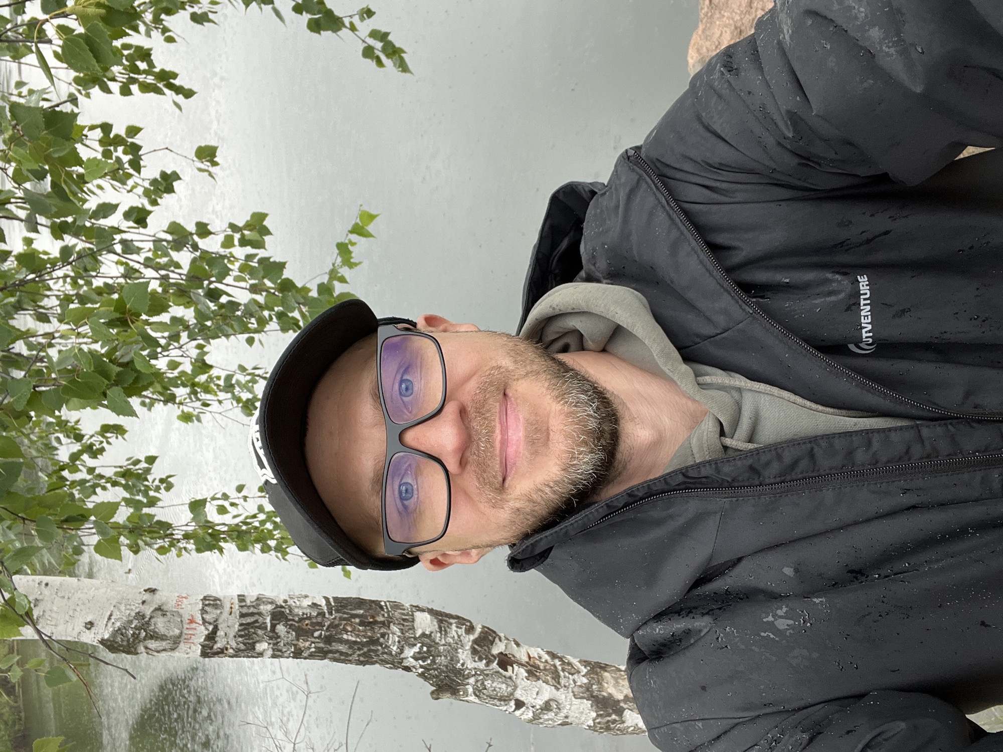 A man wearing a black baseball cap, black-rimmed glasses, and a black waterproof jacket over a grey hoodie takes a selfie outdoors. He has a beard and is smiling. A birch tree with markings on its trunk stands to his left, and green leaves hang from branches above him. The background is a grey, overcast body of water. The scene appears to be in a park or natural area, possibly near Chelyabinsk, Russia, given the presence of birch trees and the overcast, cool weather. The jacket appears wet, suggesting it may be raining or has recently rained. The text "OUTVENTURE" is visible on the lower left side of the man's jacket.