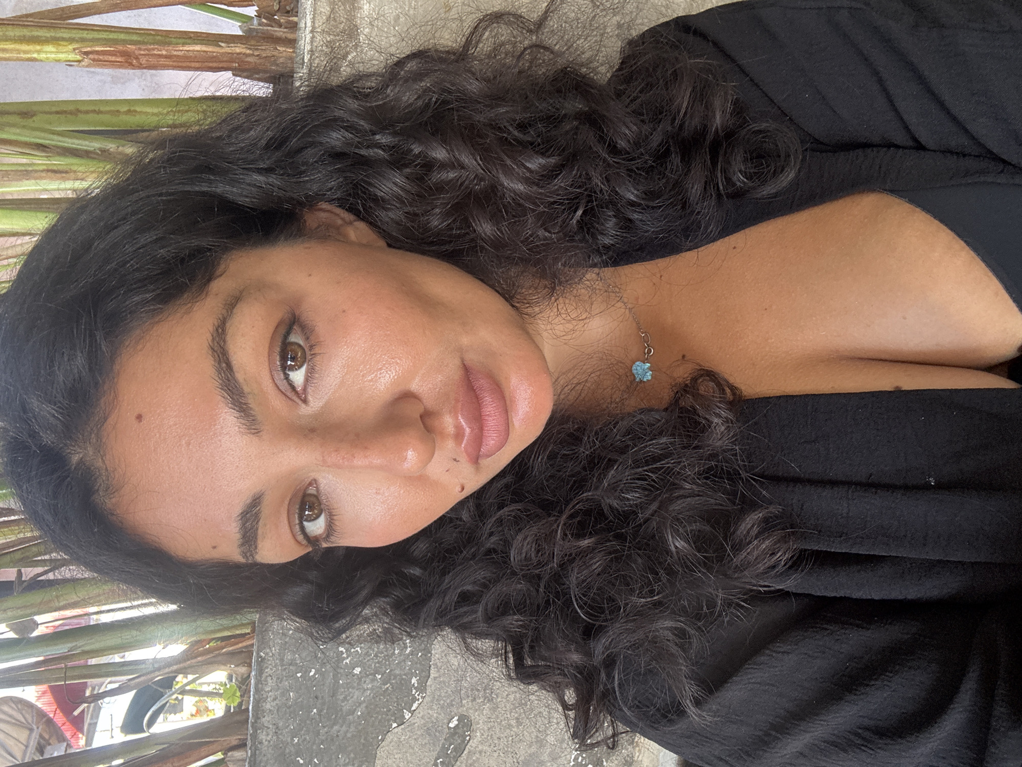 A woman with brown hair and brown eyes is looking at the camera. She is wearing a black shirt and a necklace with a blue pendant. The background includes a concrete surface and some green plants. The location is not explicitly specified as Kuta Selatan, Indonesia, although it could be in the general area.
