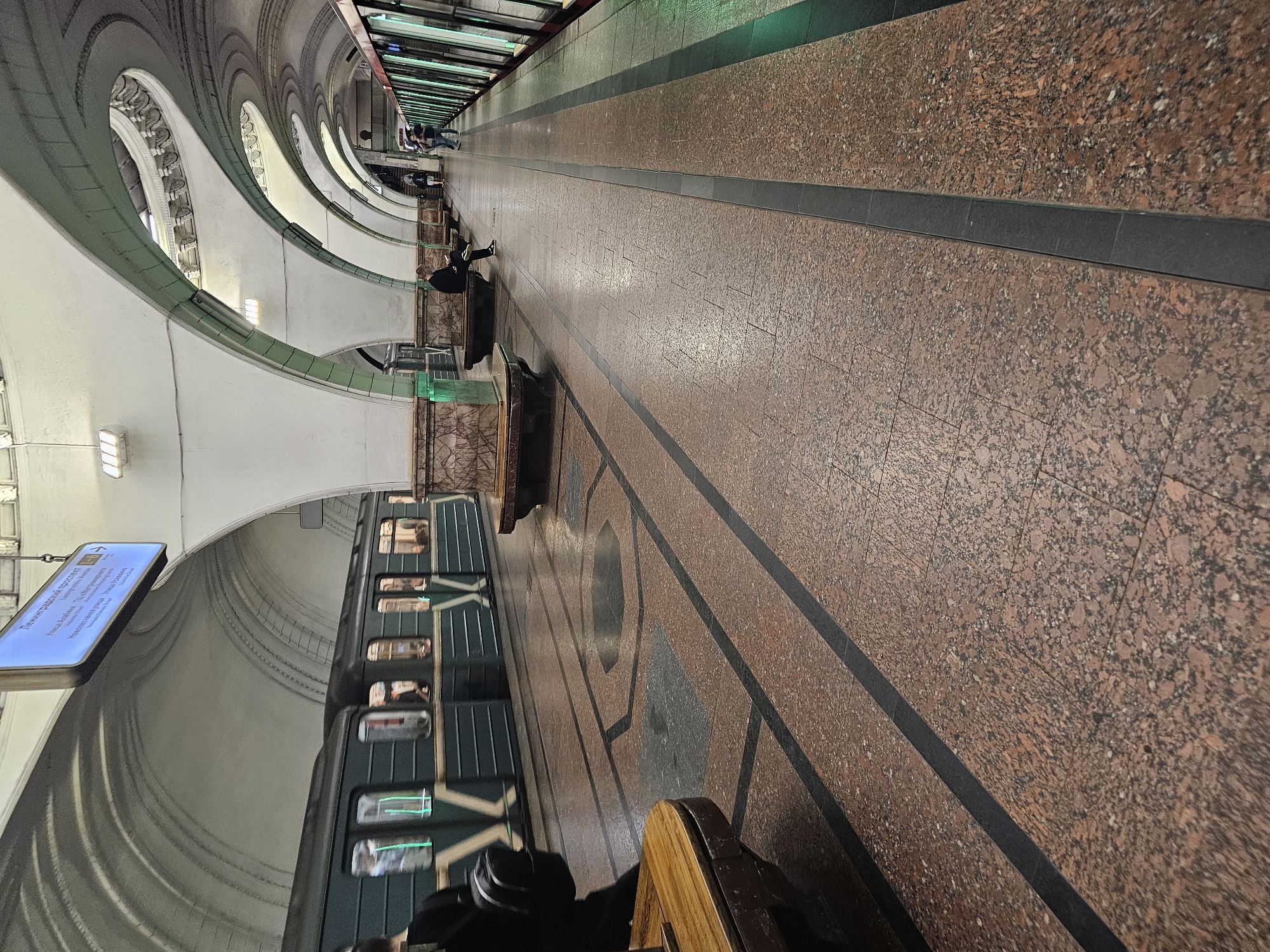 This image depicts an interior view of a Moscow Metro station, likely underground given the architecture and lighting. The scene is characterized by a long, curved platform with a train stopped alongside it. The platform floor is paved with large, reddish-brown granite tiles with dark grey inlays.
Several individuals are visible in the station. One person is seated on a bench on the platform, facing away from the camera. Further down the platform, other figures can be seen standing and sitting, some near the edge of the platform. A train, with its windows reflecting the station's lights and interiors, is stationary. The train cars are dark green.
The station's architecture is a prominent feature. The ceiling is a series of white arches, with decorative elements and green trim along the edges. The overall impression is one of grandeur and a somewhat dated but well-maintained public space. There are some lights visible on the ceiling. On the left side of the platform, there is a blue digital display board with text that appears to be in Cyrillic, likely providing information about train schedules or station services.
The lighting suggests it is not daytime, and the weather is irrelevant as the scene is indoors. The atmosphere appears calm and ordinary, typical of a functioning metro station. The image has been rotated, showing the platform curving to the left.