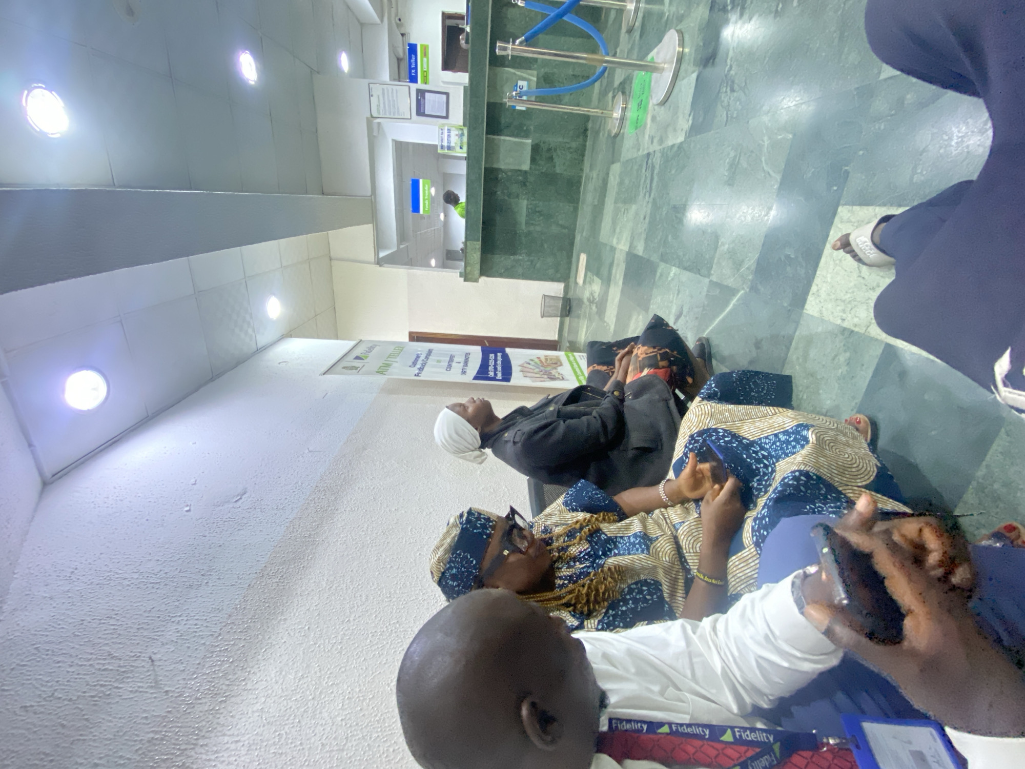 In an indoor scene that appears to be a bank or financial institution in Jos, Nigeria, three individuals are seated in a waiting area. On the left, a person wearing a white shirt and a red lanyard with "Fidelity" printed on it is looking down at a mobile phone. In the center, a person with braided hair, wearing a patterned blue and gold dress and glasses, is also looking at their phone. To the right, another person, wearing a dark jacket and a white headscarf, is seated and looking forward. The environment features a textured white wall, a drop ceiling with lights, and a floor with a green marble pattern. A counter with a green marble finish is visible in the background, with a staff member in a bright green shirt attending to it. Blue stanchions with ropes are set up in front of the counter, indicating a queueing system. Posters and signage are present on the walls.