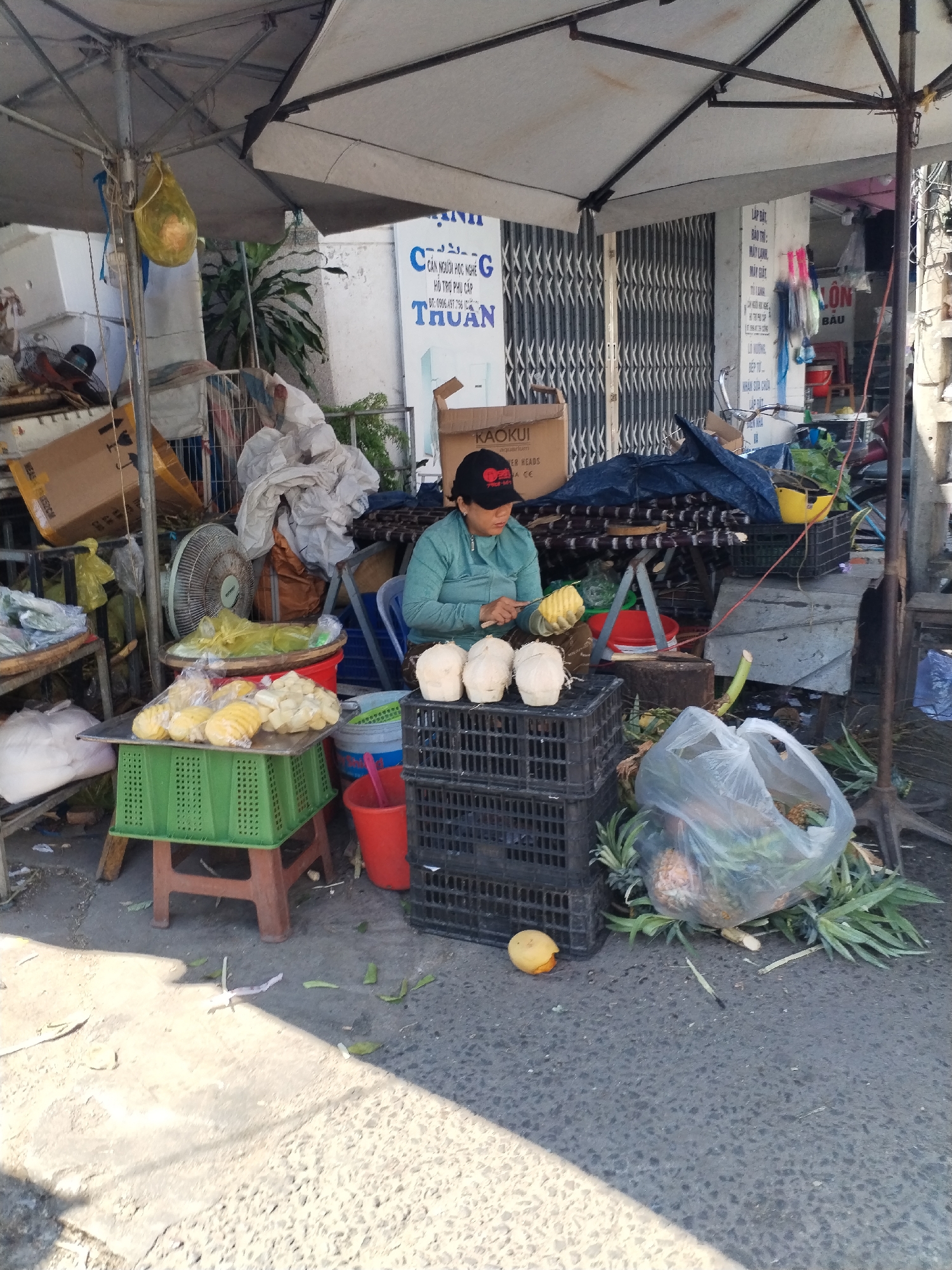 A woman sits behind stacked black crates under a large white umbrella, preparing fruit for sale on a street in Bac Nha Trang Ward, Vietnam. She wears a green long-sleeved shirt and a black hat and is cutting a yellow fruit with a knife. Three white coconuts rest on the crates beside her. To her left, yellow chunks of fruit, some in plastic bags, are arranged on a green crate atop a small stool. A plastic bag filled with pineapples and pineapple leaves sits on the ground next to the crates. A peeled mango lies on the ground nearby. Debris and tools litter the scene. In the background, the façade of a building displays Vietnamese text.