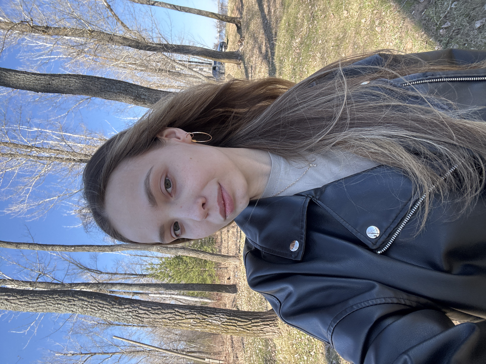 A young woman is taking a selfie outdoors, likely in a park or wooded area in Chaikovsky, Russia. It is daytime, with a clear blue sky. The trees are mostly bare, suggesting early spring. The woman is wearing a black leather jacket and a beige shirt. She is wearing gold hoop earrings and a necklace. The ground is covered in grass and some dead leaves.