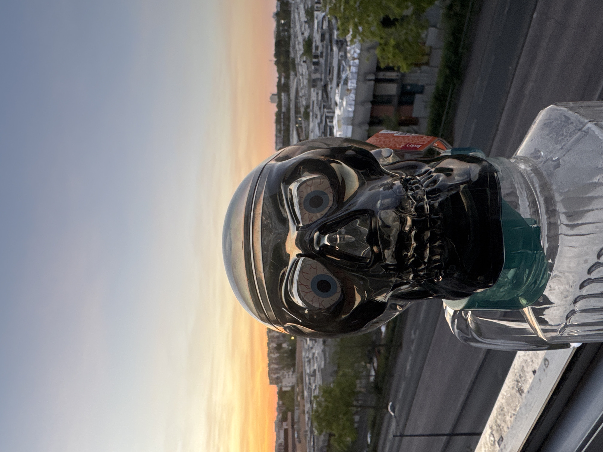 A close-up, eye-level, outdoor, vertical medium shot captures a reflective, chrome skull sculpture with visible blue-veined eyeballs, set against a hazy sunset sky above a cityscape. The skull, which appears to be made of glass or polished metal, is positioned in the foreground, slightly off-center to the left. A green cloth or ribbon is tied around its neck, and the skull rests on top of a clear, ribbed glass container that extends out of frame at the bottom. A red tag with white lettering is partially visible on the right side of the skull's neck. In the background, a wide, empty road runs horizontally across the frame, bordered by trees and a dense collection of buildings with flat roofs. The sky transitions from a pale blue at the top to a warm orange and yellow gradient near the horizon, suggesting dusk. The overall scene evokes a slightly surreal or artistic ambiance.