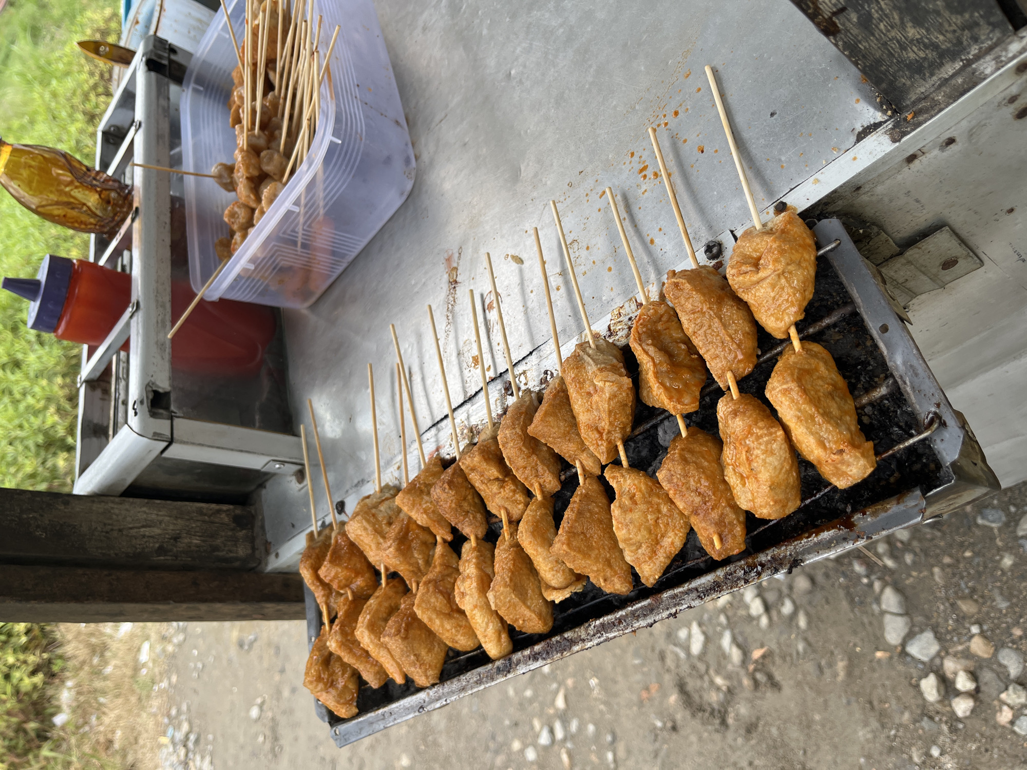 This image captures an outdoor street food stall in Matangpanyang, Indonesia, displaying various skewered items for sale. The primary focus is a rectangular metal charcoal grill rack on the left, holding 25 wooden skewers arranged in five rows of five. Each skewer holds one or more pieces of light orange-brown, irregularly shaped, grilled food, consistent with fried tofu (tahu goreng) or a similar fried snack, coated in a reddish seasoning or sauce. The grill rack itself shows signs of heavy use, with black charring and some remnants of drippings.
To the right of the grill, on a metallic countertop, a clear rectangular plastic container holds a quantity of smaller, roundish, light-colored skewered items, consistent with meatballs or fish balls (bakso), also on wooden skewers. These items are not currently on the grill but are prepared for cooking or serving.
Behind the container, a metal rack holds two bottles: a red plastic squeeze bottle with a purple cap, typically used for chili sauce or ketchup, and an inverted, clear brown bottle, likely containing soy sauce or another liquid condiment.
The stall appears to be a basic, mobile setup, with a dark wooden vertical support beam visible on the left. The ground immediately surrounding the stall is unpaved, consisting of dirt mixed with small grey and white pebbles and rocks. In the background, beyond the stall, dense green foliage is visible under natural light, indicating an outdoor, possibly semi-rural or roadside, setting. No individuals are visible in the frame.