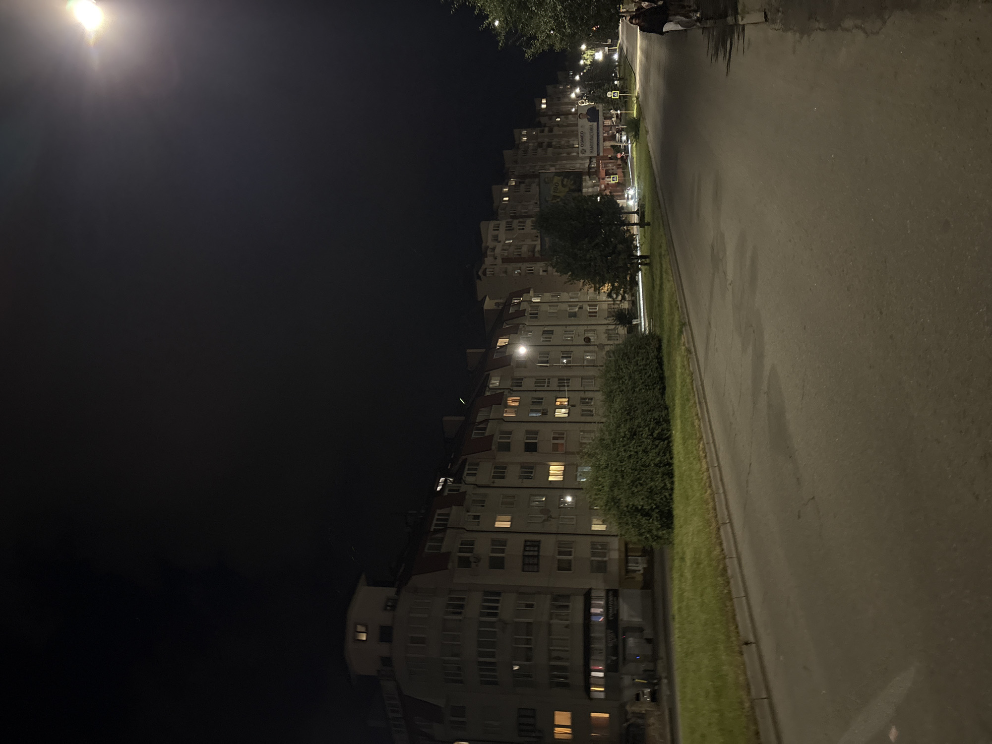The image captures a nighttime view of a street in Ternopil, Ukraine, likely along a residential area. A long, multi-story apartment building dominates the left side of the frame, its windows illuminated with a warm, yellow light, suggesting occupancy. In front of the building, a strip of green grass and a dense hedge line the sidewalk. The street itself is paved and appears to be empty of major traffic, with only faint reflections on its surface.
Further down the street, the apartment building continues, with more illuminated windows. Several streetlights are visible, casting pools of light onto the pavement and illuminating trees and other vegetation along the roadside. A few signs are visible on the buildings and in the distance, one of which appears to be a billboard with images and text.
On the right side of the image, a sidewalk runs alongside the street, and two individuals are walking away from the camera. Their figures are dark against the illuminated street. The overall atmosphere is quiet and urban, characteristic of a residential district at night. The sky is dark, with a bright light source, possibly a streetlamp or a distant light, visible in the upper right corner, creating a lens flare effect.