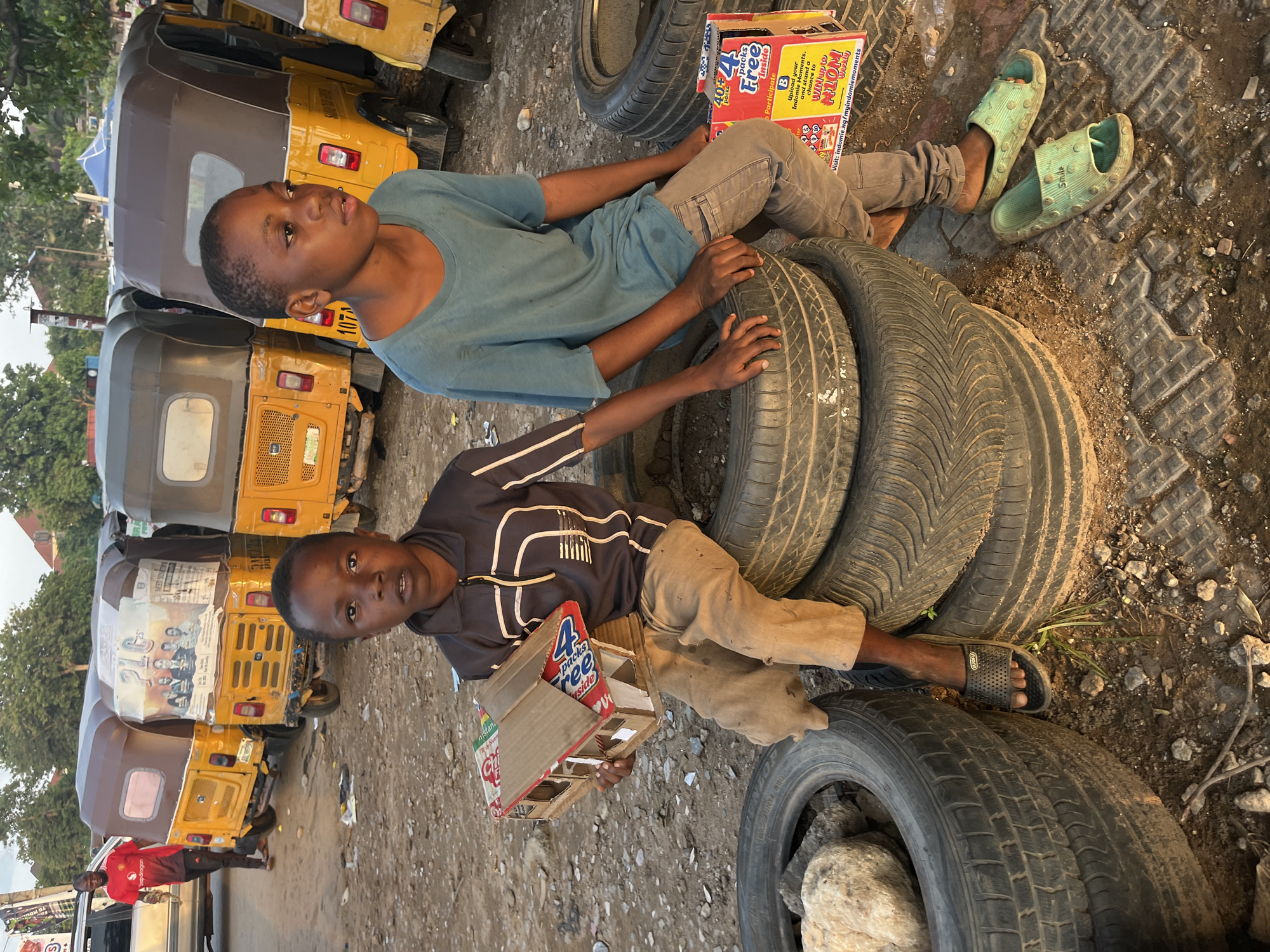 Two young boys sit on stacks of old tires in what appears to be an outdoor informal market or roadside area in Abuja, Nigeria. The boy on the left, wearing a dark, zip-up jacket with a white stripe design and khaki pants, holds a small, handmade cardboard house. The boy on the right, dressed in a light blue t-shirt and gray pants, looks off to the side.
In the background, several yellow auto-rickshaws, commonly known as "Keke NAPEP" in Nigeria, are parked. One of the rickshaws has a large poster on its rear featuring a group of people, possibly politicians, with the text "PLC" visible. The ground is dirt and gravel, scattered with debris. The overall scene suggests an informal, possibly impoverished, urban environment. The lighting indicates it is daytime, likely late afternoon, with the sun casting long shadows.