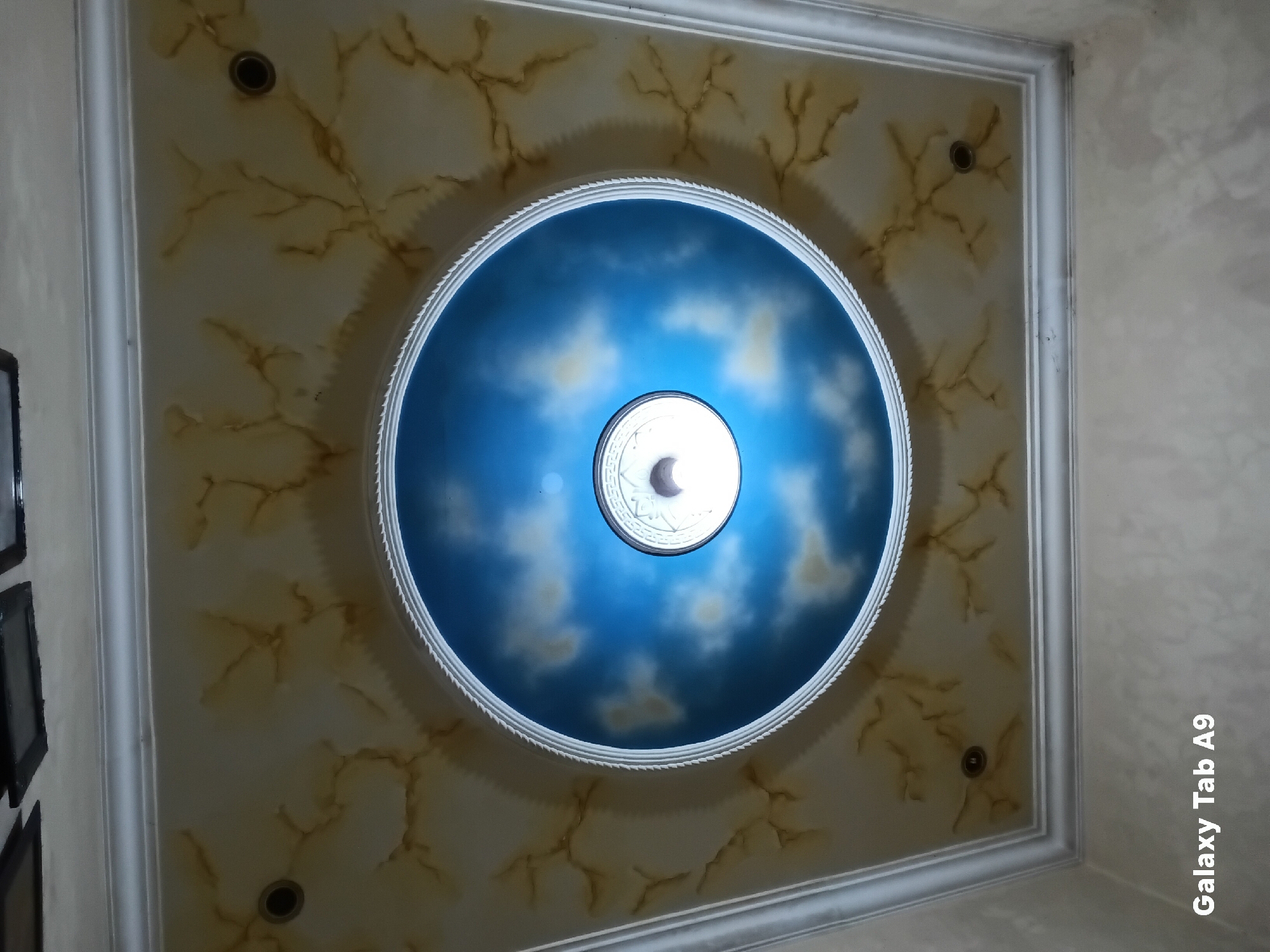 The image shows a ceiling with a decorative design. The ceiling is a square shape with an ornate, circular design in the center. The circular design depicts a blue sky with clouds, and a light fixture is visible in the center of the circle. The outer part of the ceiling has a faux marble or textured pattern in beige and brown tones, with decorative trim along the edges of the square. The overall impression is one of an interior space, possibly a room, with a focus on decorative ceiling treatment. The image was captured on a Samsung Galaxy Tab A9. There is no information in the image to suggest a specific location in Trenggalek, Indonesia.