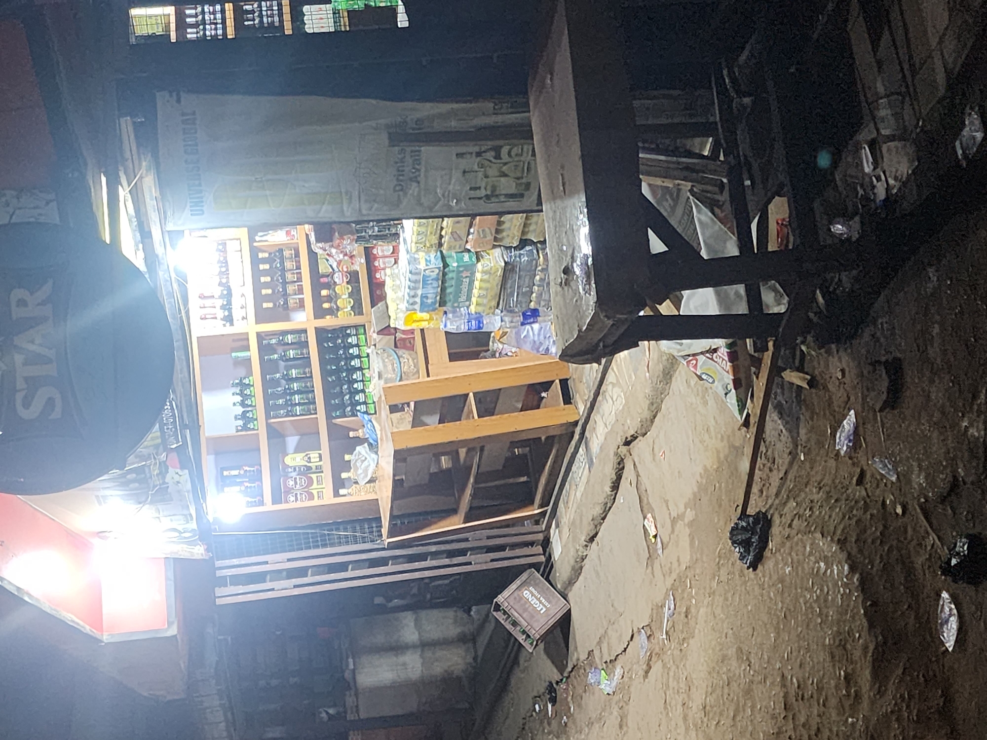 The image shows a street scene at night in Obiaruku, Nigeria. The location appears to be a small commercial area. A store is visible with shelves stocked with drinks, including bottled water and alcoholic beverages. A banner reading "UNIVERSE GLOBAL" and "Drinks Avail..." hangs above the store. A "STAR" sign is visible on the left. The ground is littered with debris and trash. A wooden table and various metal structures are present in the foreground, partially obstructing the view. A crate labeled "LEGEND" is lying on the ground. No people are clearly visible. The lighting is uneven, with bright areas near the store and dark shadows elsewhere.