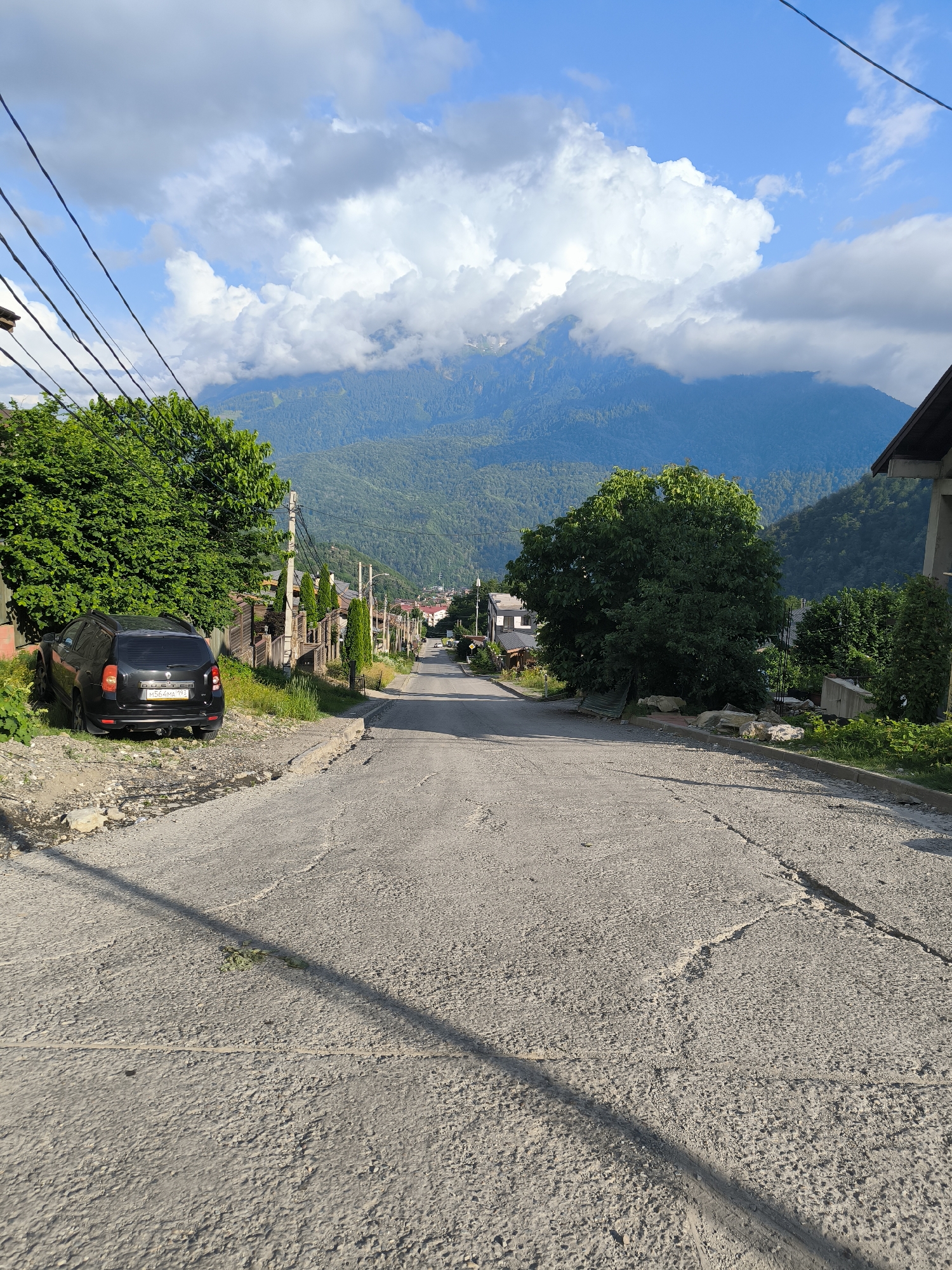 This is a vertical, eye-level, outdoor, full shot of a winding, paved road in a mountainous, rural setting. The road appears to be well-worn and cracked, with a gravel shoulder on the left side. A black Dacia Duster is parked on the gravel shoulder to the left, with its license plate visible, reading "M564MA 199".
The road leads uphill, disappearing into the distance. On either side of the road are houses, some appearing to be residential, and mature trees. The overall impression is a quiet, residential street in a mountainous village.
In the background, lush green mountains rise, partially obscured by dramatic, white cumulus clouds that are tinged with blue from the sky. The sunlight suggests it is daytime, likely late morning or early afternoon, with fair weather. The scene is peaceful and serene, evoking a sense of calm and natural beauty. The setting appears to be Krasnaya Polyana, Russia, a well-known mountain resort area. No people are visible in the image.