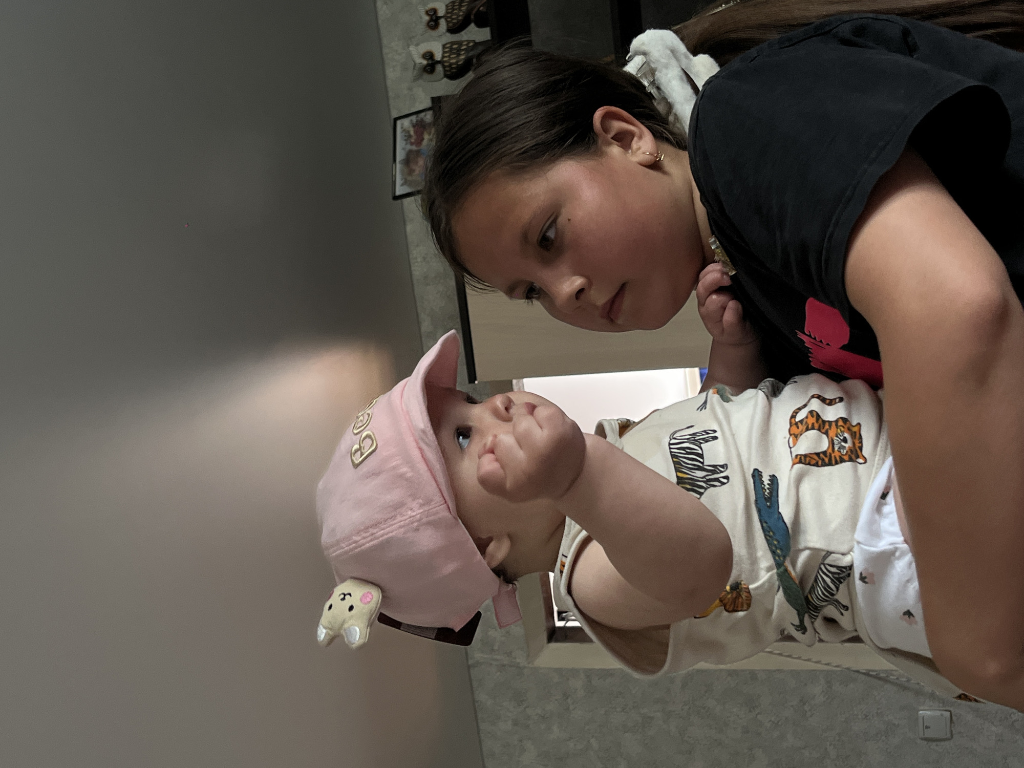 A child wearing a black T-shirt looks at a baby wearing a pink cap and clothes with animal prints. The location is Novopokrovskaya, Russia.