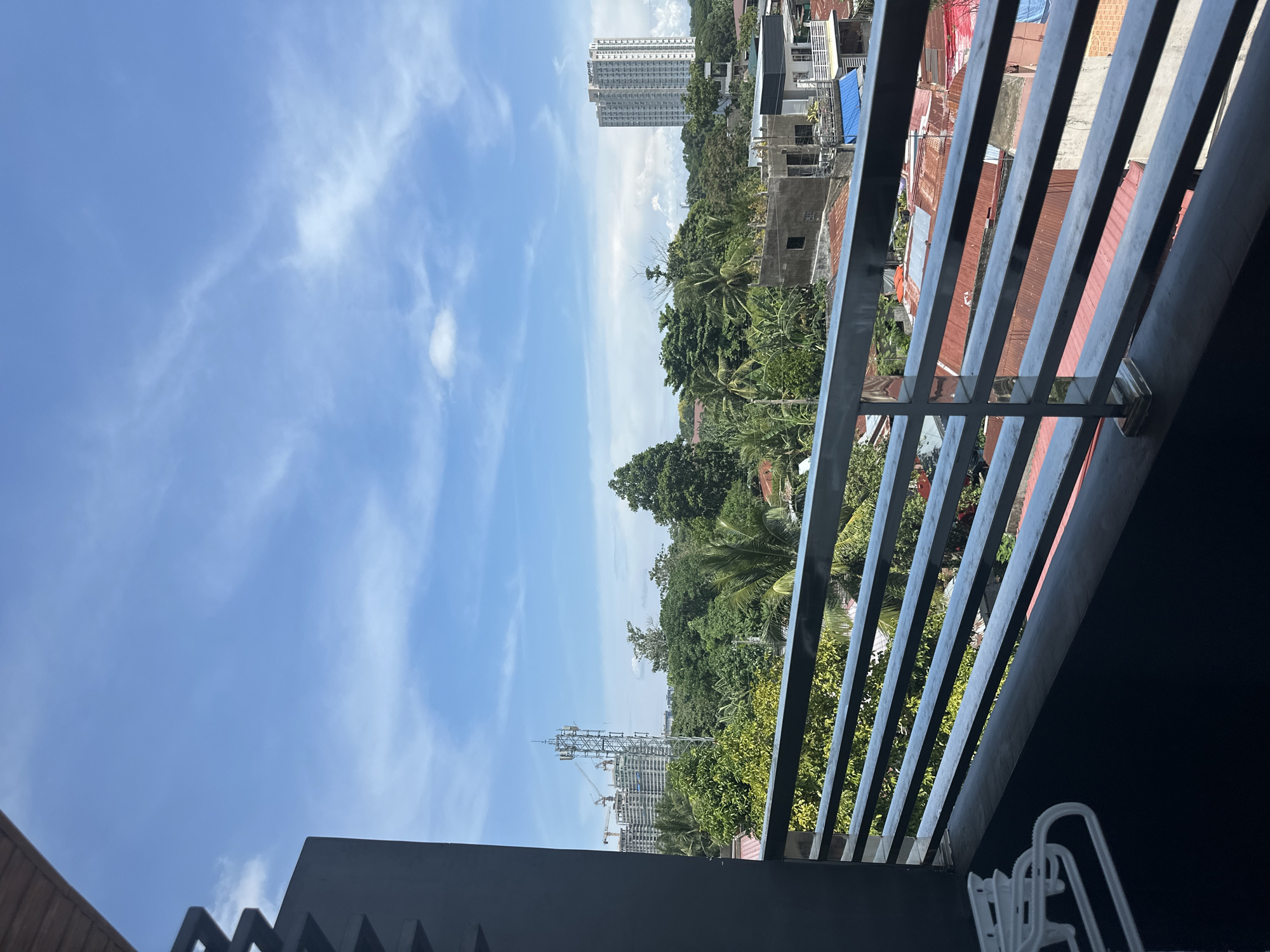 The image is taken from a high vantage point, possibly a balcony, in Davao City, Philippines. The sky is blue with scattered clouds. The view is partially obstructed by a gray metal railing. Beyond the railing, there are buildings, lush green trees, and residential structures with red and blue corrugated iron roofs. In the distance, there is a tall high-rise building. To the left, a construction site with a tower crane and incomplete structure are visible. The overall scene depicts a mix of urban development and natural landscape in a tropical environment.