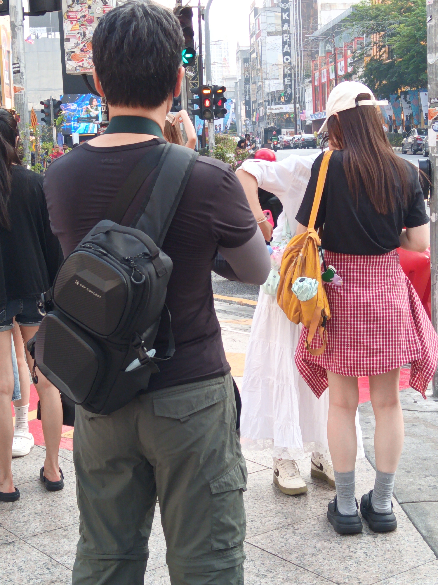 The image captures a street scene in Bukit Bintang, Kuala Lumpur, Malaysia. In the foreground, a person wearing a black t-shirt and green cargo pants is viewed from behind, carrying a black sling bag. To their right, a woman in a white dress and a person in a black t-shirt and a red and white checkered skirt stand together. The woman in the white dress has a yellow bag with items hanging from it. In the background, the street is lined with buildings, including a "KARAOKE" sign, and traffic. Traffic lights are visible, indicating an intersection. Several people are walking on the sidewalk. The overall atmosphere appears to be that of a busy urban commercial district.
