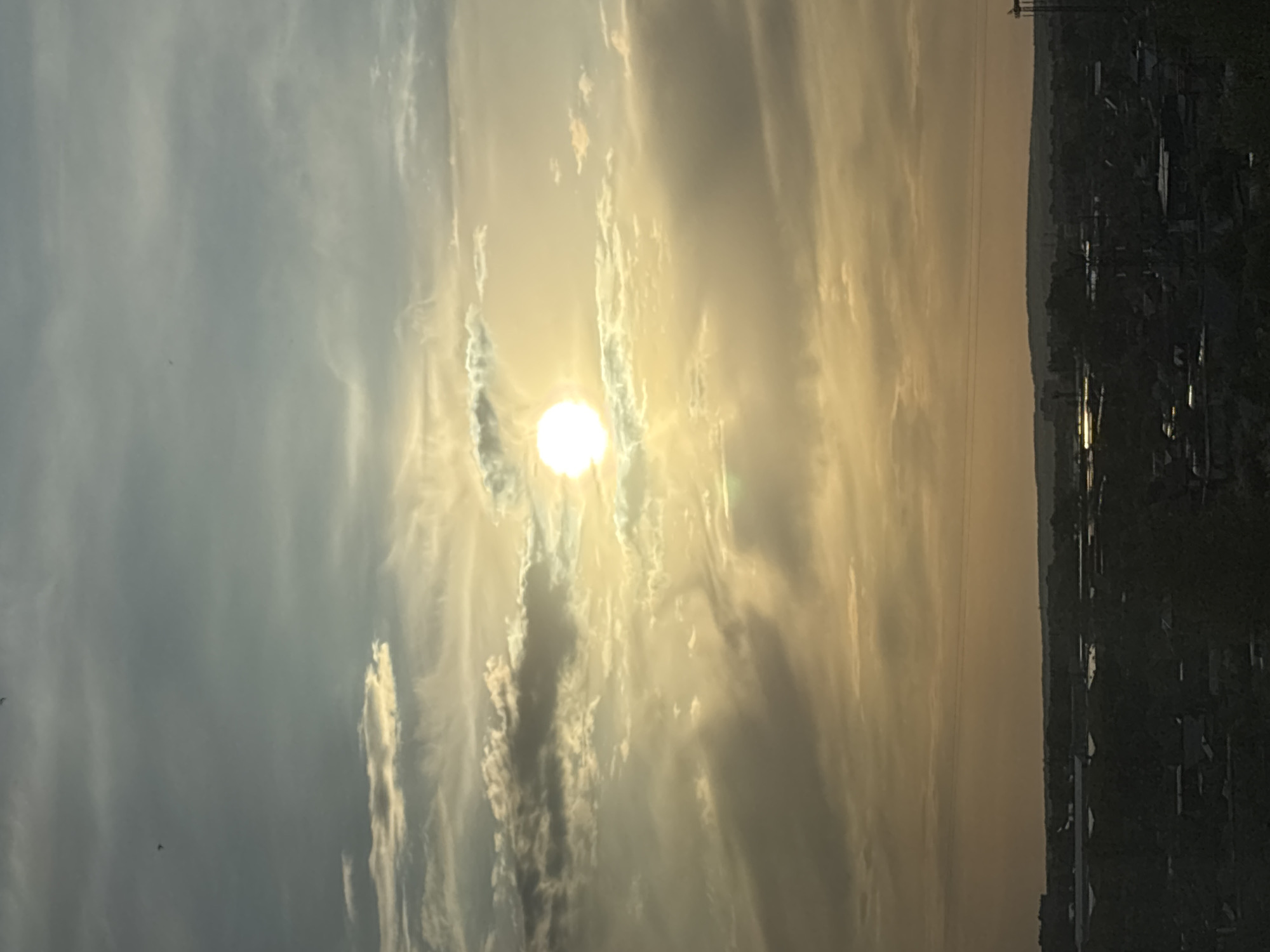 The image depicts a sunset in Chaikovsky, Russia. The sun, a bright white orb, is partially obscured by striated clouds, creating rays of light. The sky transitions from blue-grey in the upper left to a warm golden hue where the sun is. The silhouette of buildings and trees is visible along the right edge of the frame, indicating an urban or suburban area. Power lines can be seen cutting through the sky near the skyline. The scene suggests a serene, natural setting juxtaposed with human-made structures.