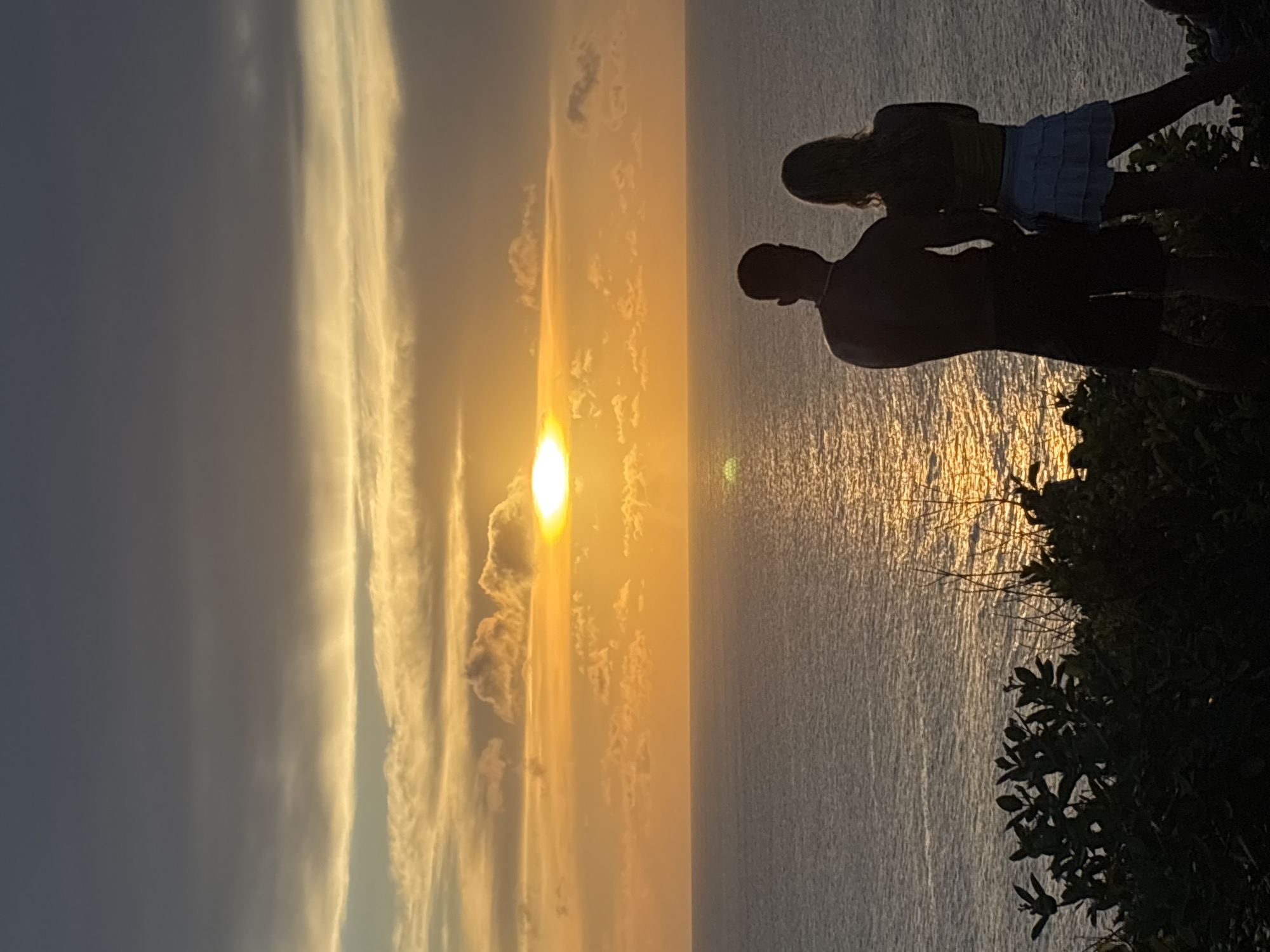 Captured at Uluwatu, Indonesia, the image portrays a sunset viewed from a high vantage point, likely a cliff. To the right are two people silhouetted against the sunlit ocean. The taller figure, possibly male, is beside the slightly shorter figure, wearing a light-colored, short skirt. Dense foliage frames the right side of the image. The ocean reflects the golden light of the setting sun, creating a bright path across the water. To the left, the sky transitions from deep blue to a mix of golden hues and cloud formations around the setting sun.