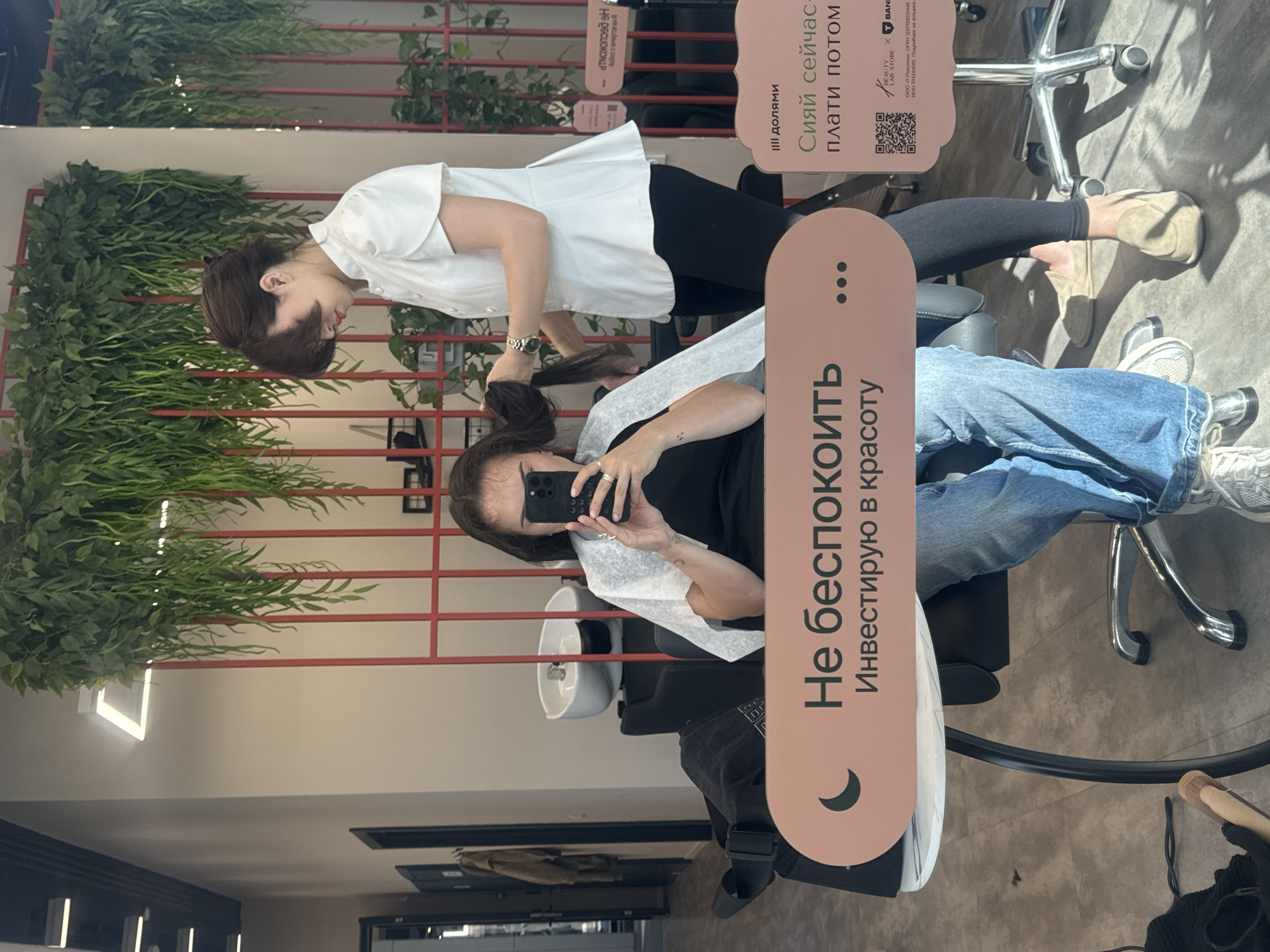This image captures a moment in a modern hair salon, presented as a mirror selfie taken by a client. Two women are visible: a client seated in a salon chair and a stylist attending to her hair.
The client, with dark hair, is wearing a black top or dress and blue jeans, covered by a white disposable cape. She is holding a black smartphone, reflecting her image in the mirror, indicating she is taking the photo. Her expression is not clearly visible, but she appears relaxed.
The stylist, standing behind the client, has dark brown hair and is dressed in a white short-sleeved shirt and dark pants. She is actively working on the client's hair.
The salon interior appears bright and clean, with decorative elements including green artificial plants arranged on a red grid structure, giving the space a contemporary aesthetic. A white wash basin is partially visible behind the client.
Two vertical signs with text are prominent. The larger sign reads: "Не беспокоить" (Do not disturb) and "Инвестирую в красоту" (Investing in beauty). Above it, a smaller sign advertises a payment service: "III долями" (In installments), "Сияй сейчас- плати потом" (Shine now - pay later). This sign also includes a QR code and fine print mentioning "BEAUTY STORE x BANK" and details of a company named "ООО «Т-Покупки»," including its OGRN and INN numbers, and a reference to "dolyami" (installments). The context suggests a beauty treatment taking place during the day.