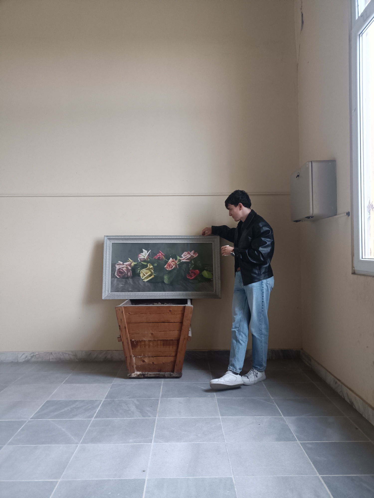 An individual wearing a black leather jacket, light blue jeans, and white sneakers stands inside a room with pale yellow walls and a gray tiled floor. A painting depicting roses in muted colors rests on top of a wooden crate or container. The person appears to be adjusting or examining the painting. A silver electrical box is mounted on the wall near a window with a white frame. The setting appears to be an interior space within a building, possibly in Plovdiv, Bulgaria, given the architectural style and tiled floor reminiscent of Eastern European interiors. The image appears candid, capturing a moment of interaction between the person and the artwork.