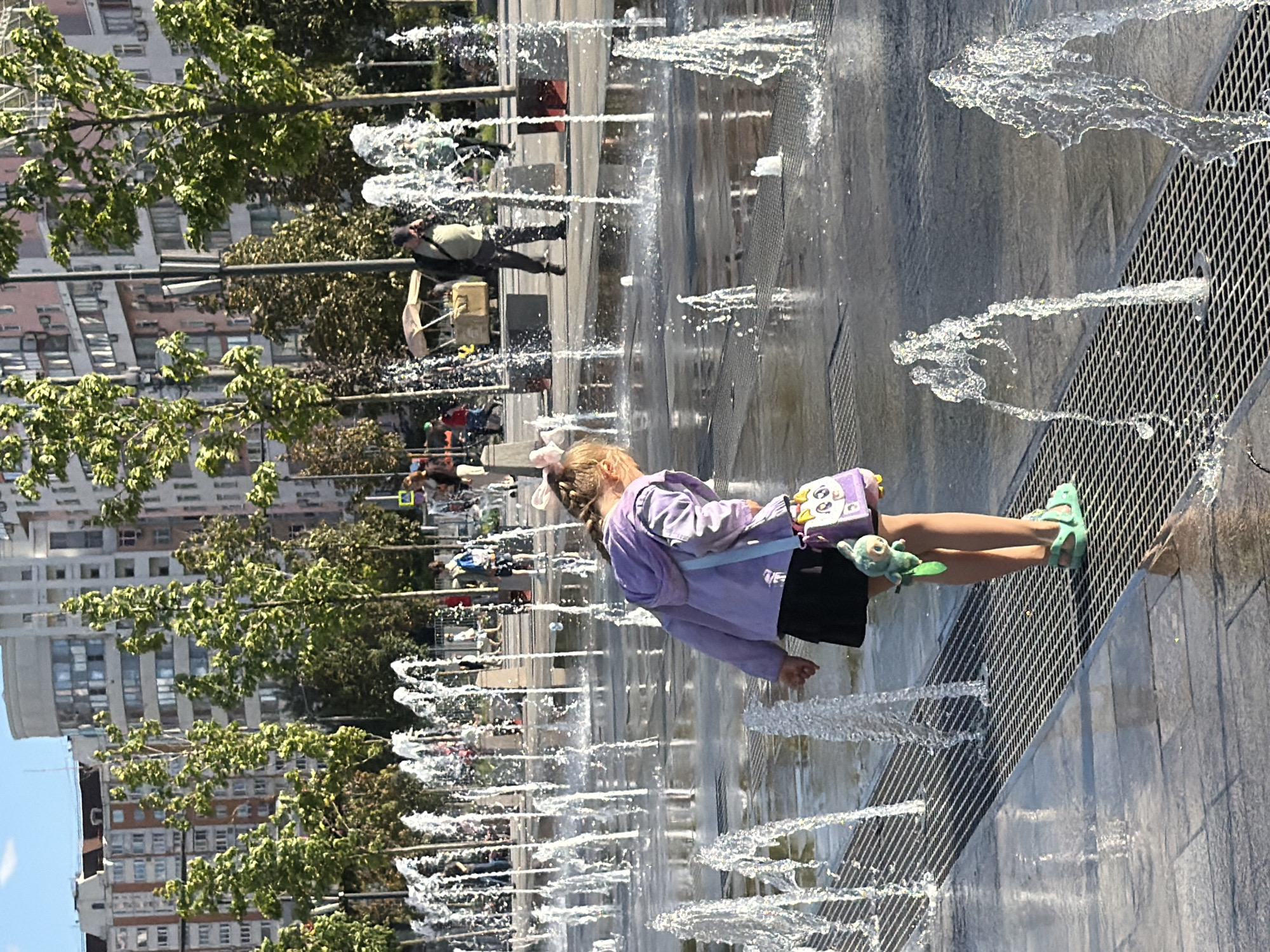 In an outdoor urban setting, a young girl is shown playing in a splash pad. She is wearing a purple hoodie, a black skirt, and green sandals. A small stuffed animal is attached to her belt, and a purple bag with cartoon eyes is slung over her shoulder. She is standing on a metal grate as water sprays around her. The splash pad is surrounded by trees and tall buildings, suggesting a city park or public plaza. Several other people are visible in the background, some walking and others observing the fountain. The sun appears to be shining, casting reflections on the wet ground and water. The overall atmosphere seems lively and summery.