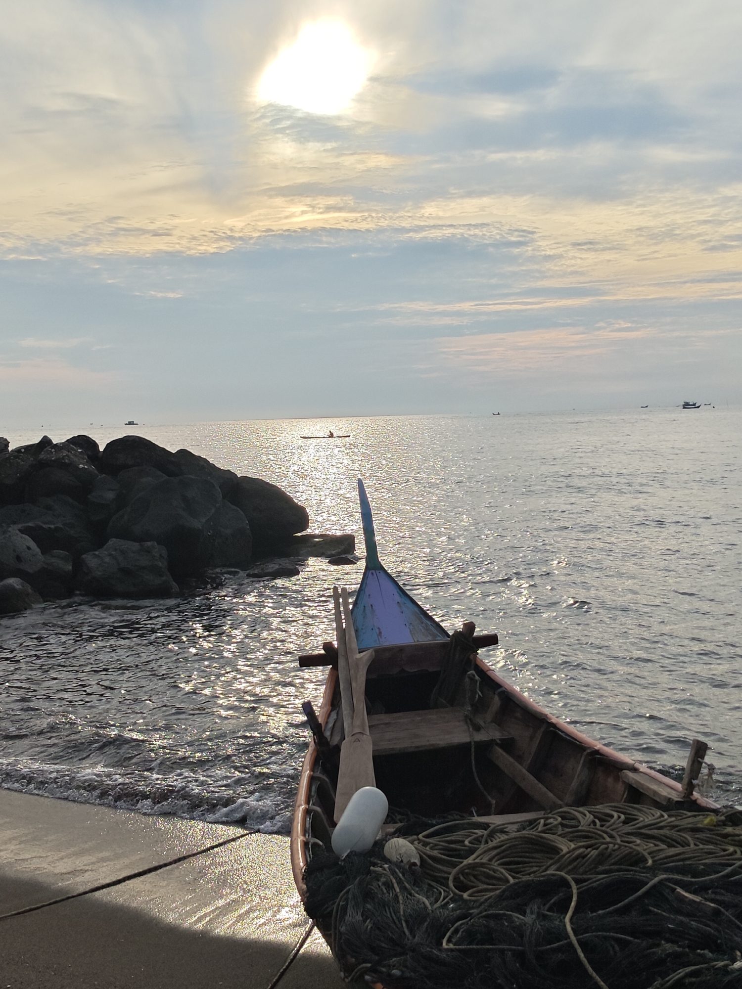 This image depicts a serene coastal scene, likely at sunrise or sunset, near the city of Lhokseumawe, Indonesia. A traditional wooden fishing boat rests on the sandy shore, its bow pointing towards the water. The boat is laden with fishing nets and ropes, suggesting its purpose. In the background, the calm sea reflects the warm glow of the sun, creating a shimmering effect on the water's surface. Several other small boats and what appears to be a distant landmass or larger vessel are visible on the horizon. To the left of the frame, a cluster of large, dark rocks forms a natural breakwater, with gentle waves lapping at the shore. The sky above is a soft blend of blue and pale yellow, with scattered clouds that are illuminated by the sun. The overall atmosphere is peaceful and tranquil.