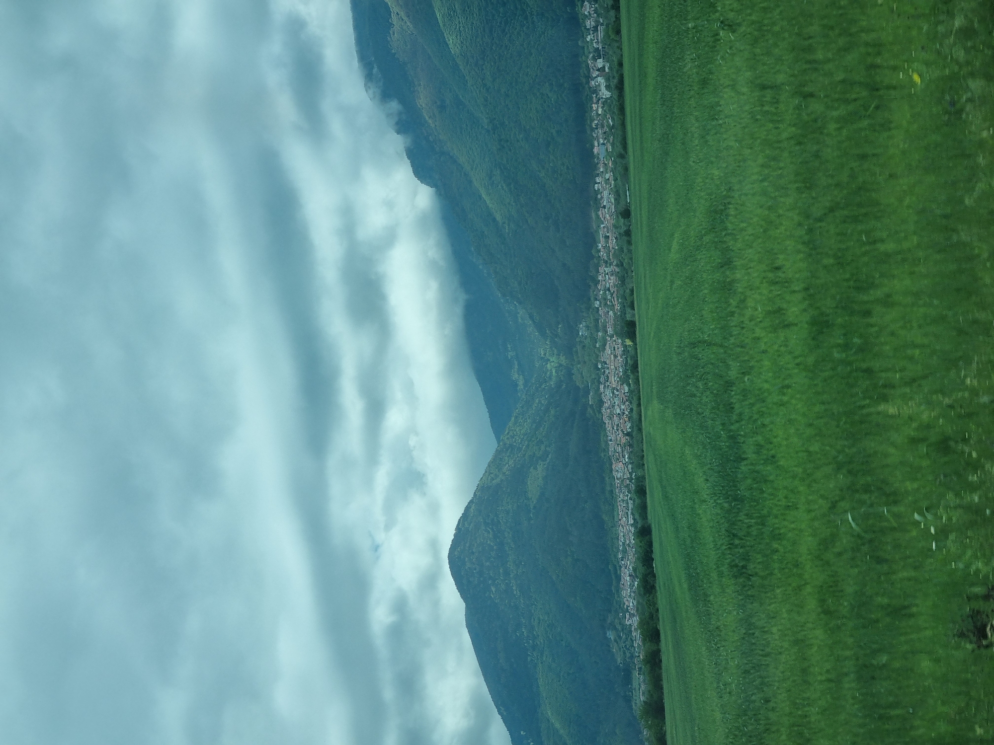 The image shows a landscape in Yahinovo, Bulgaria. Mountains covered with lush green vegetation rise from the horizon, their slopes partially obscured by clouds. A town nestles at the foot of the mountains, its buildings stretching along the base. To the right, a vast expanse of green field extends into the foreground. The sky is overcast with thick, layered clouds.