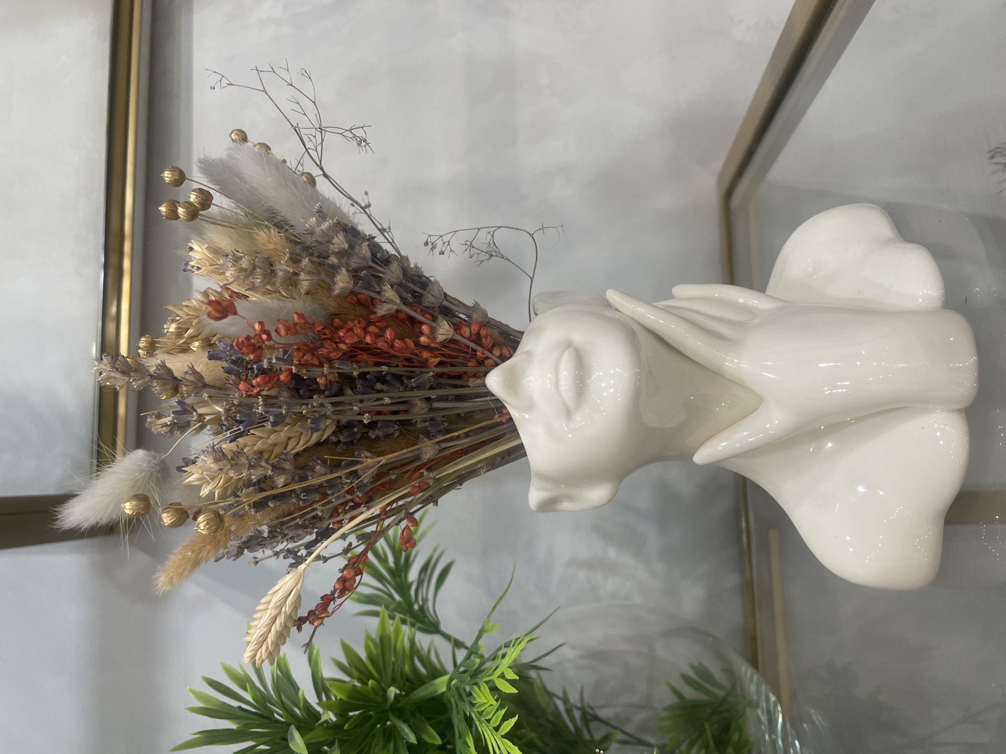 The image shows a white ceramic bust of a woman holding her chin, with a bouquet of dried flowers in her head. The bust is placed on a glass shelf with a golden frame in the background. To the left of the bust, there is a green potted plant. The background is a light gray wall with subtle textured patterns. The scene appears to be an indoor setting, possibly a home or a shop display. There is no visible indication of the city of Saint Petersburg, Russia, or any specific geographical location. The lighting is soft and diffused, highlighting the glossy surface of the ceramic bust and the various textures of the dried flowers.