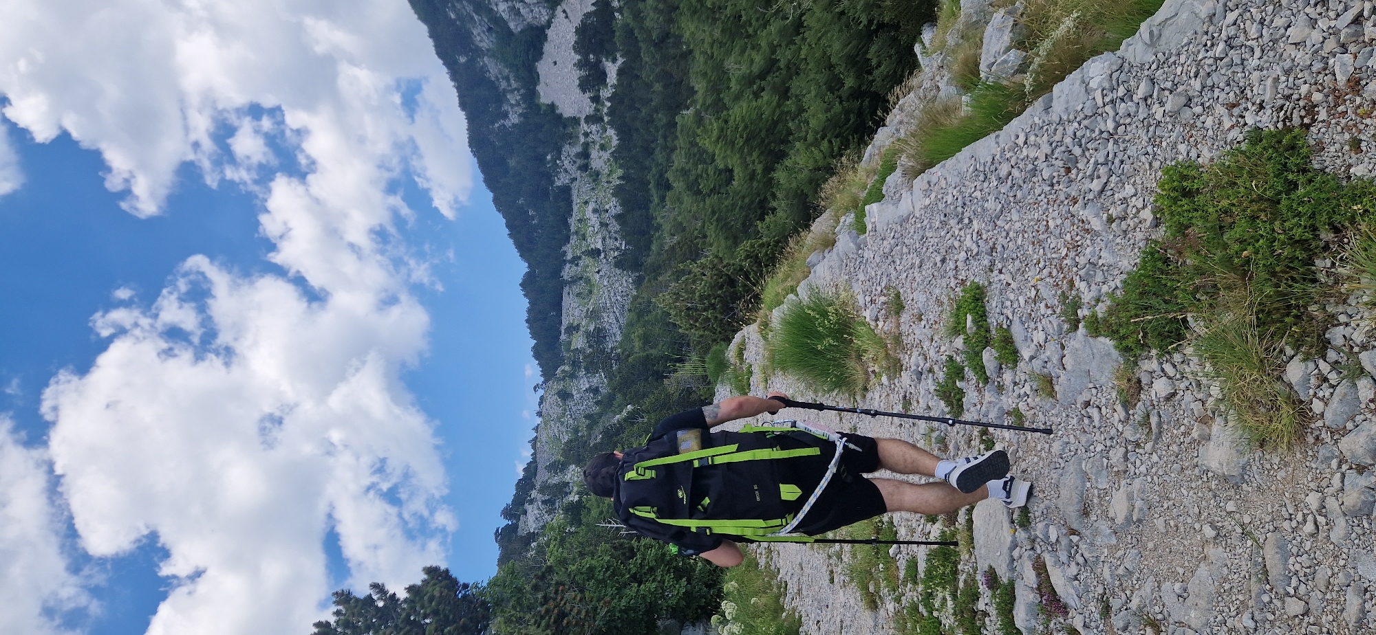A person with a black backpack with bright green straps and hiking poles is walking on a gravel path uphill, towards a mountainous area covered with trees and rocks. The sky is blue with white clouds. The person is wearing black shorts and white and black athletic shoes. The scene appears to be in a natural outdoor setting, possibly a hiking trail. The location is identified as Kruševice, Montenegro.