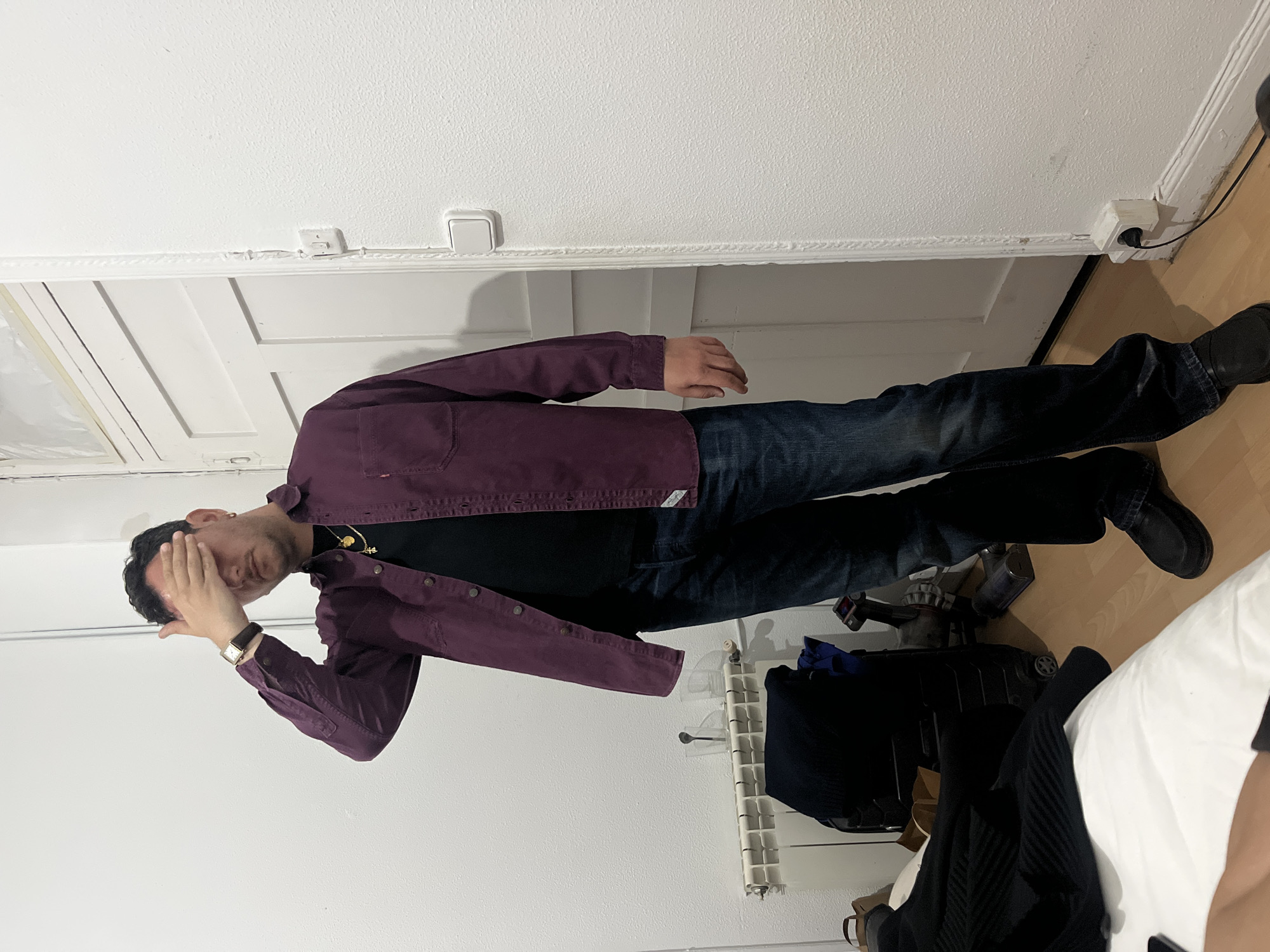 A man stands in a room with white walls and a wooden floor. He wears a purple jacket, black shirt, and blue jeans with black shoes. He covers his eyes with his left hand, wearing a watch on that wrist. There's a closed white door behind him and a radiator near his feet. A suitcase, a vacuum cleaner, and a bed with white sheets are visible in the background. The image does not show anything that indicates it is in Madrid, Spain.