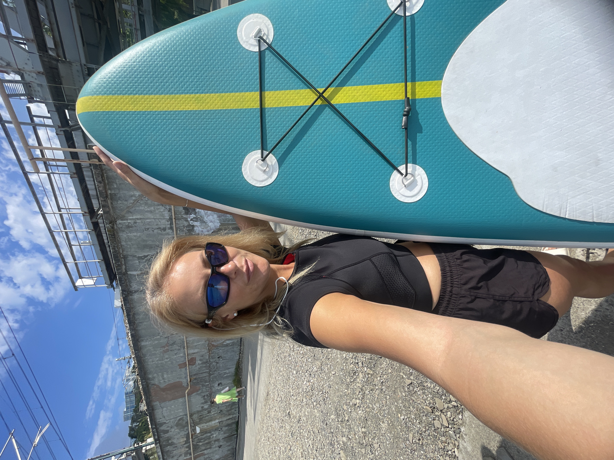 This is a portrait-oriented, vertical-format photograph of a blonde woman holding a blue and yellow stand-up paddleboard. She is wearing a black sports bra and black shorts, sunglasses, and earbuds. The paddleboard is positioned diagonally across the frame, with the woman's arm supporting it.
The setting appears to be outdoors, possibly near a body of water, given the paddleboard. In the background, there is a grey, textured wall and some industrial-looking structures made of metal beams and scaffolding. A bright blue sky with scattered white clouds is visible above. The ground is covered in gravel. There are some power lines and distant buildings in the far background. A small, blurred figure in a green shirt can be seen in the lower left background, facing away from the camera.
The lighting suggests it is daytime, likely sunny. The woman's expression is neutral, and her gaze is directed towards the camera. The overall mood is casual and active. There is no visible text in the image.