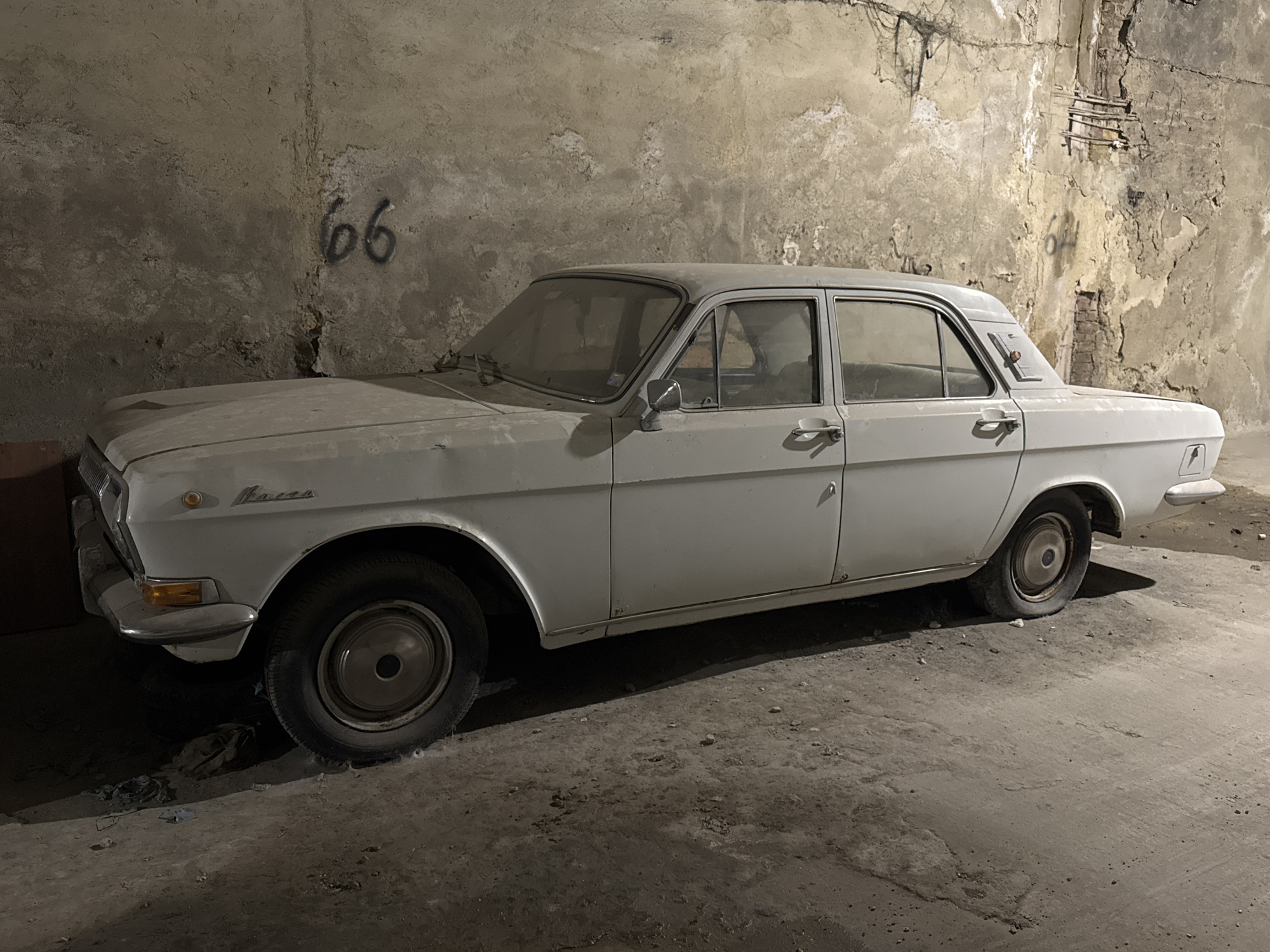 The image shows a dusty, white, four-door sedan parked indoors, possibly in a garage or abandoned building. The car appears to be a vintage model, with chrome accents on the doors and bumpers. The walls behind the car are rough and unfinished, with visible signs of wear and tear, and what appears to be the number "66" spray-painted on the wall to the left of the vehicle. The floor is concrete and shows signs of dirt and debris. The lighting is dim, suggesting an indoor, low-light environment.