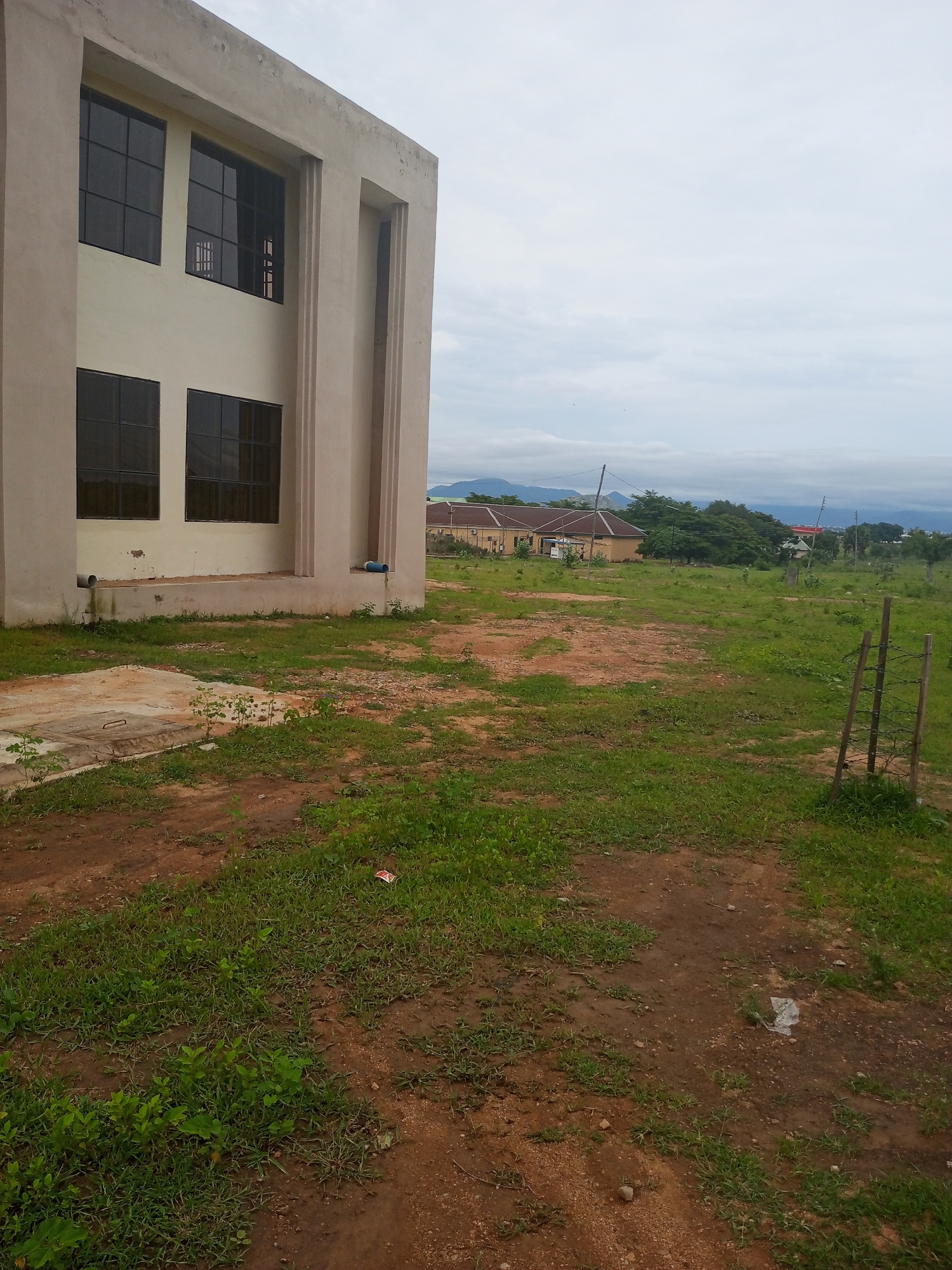 This image depicts an outdoor scene in Jalingo, Nigeria, under an overcast sky. The primary subject is a modern, pale-colored building with large, dark-paned windows. The building is partially visible on the left, with its corner and a section of its facade in view. The architectural style appears functional and somewhat stark, with vertical decorative elements on the exterior.
In front of the building, there is an area of uneven ground covered with patchy green grass and reddish-brown dirt. A concrete slab with a metal cover is visible on the left, possibly an access point for underground utilities. There are also some visible drainage pipes extending from the building. Further into the background, a row of similar buildings with light-colored walls and terracotta roofs can be seen, suggesting a larger complex or neighborhood. Lush green trees are scattered throughout the landscape, and in the far distance, mountains or hills are discernible, shrouded in mist.
The overall impression is one of a developing area or an institutional setting. The weather appears to be a typical cloudy day, with diffused light and no harsh shadows. The scene is tranquil and still, with no people or animals actively present. A small, weathered wooden fence with barbed wire is visible on the right side of the frame, adding a rustic element to the otherwise modern structures. There is also a piece of white plastic debris on the ground in the middle right of the image. No text is visible in the photograph.