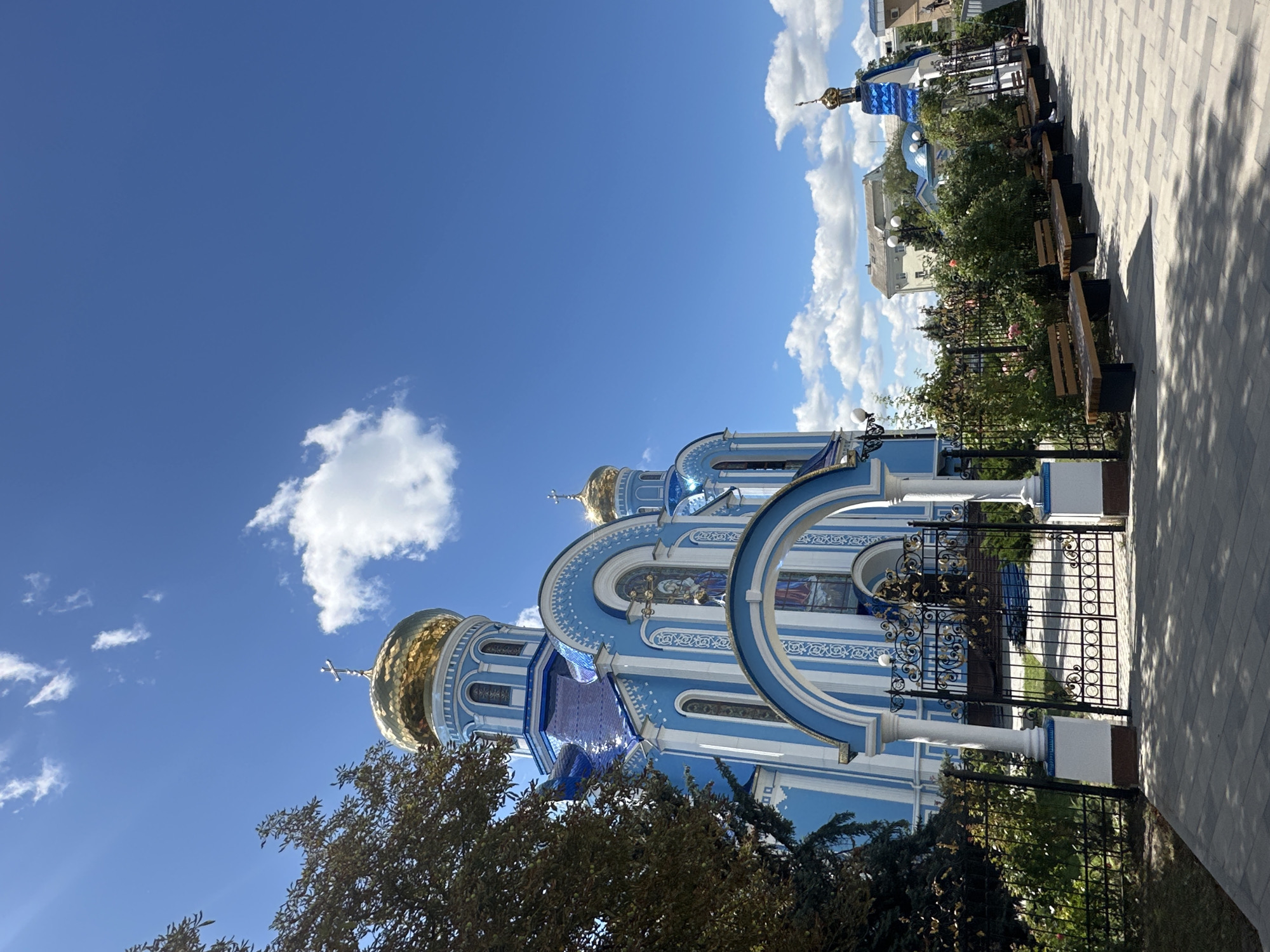 A wide shot captures the side of a predominantly blue and white Ukrainian Orthodox church with golden domes, set against a bright blue sky with scattered white clouds. The church features ornate blue roofing and white trim. In the foreground, a paved walkway leads to a decorative black metal gate adorned with gold accents, which opens to the church's entrance. To the left of the walkway, a lush green tree with dark foliage stands prominently. The right side of the walkway is lined with several wooden benches interspersed with flowering rose bushes and dense green shrubbery. In the background, other buildings and smaller blue-roofed structures are visible. The scene suggests a peaceful outdoor setting in the city of Raivka, Ukraine.