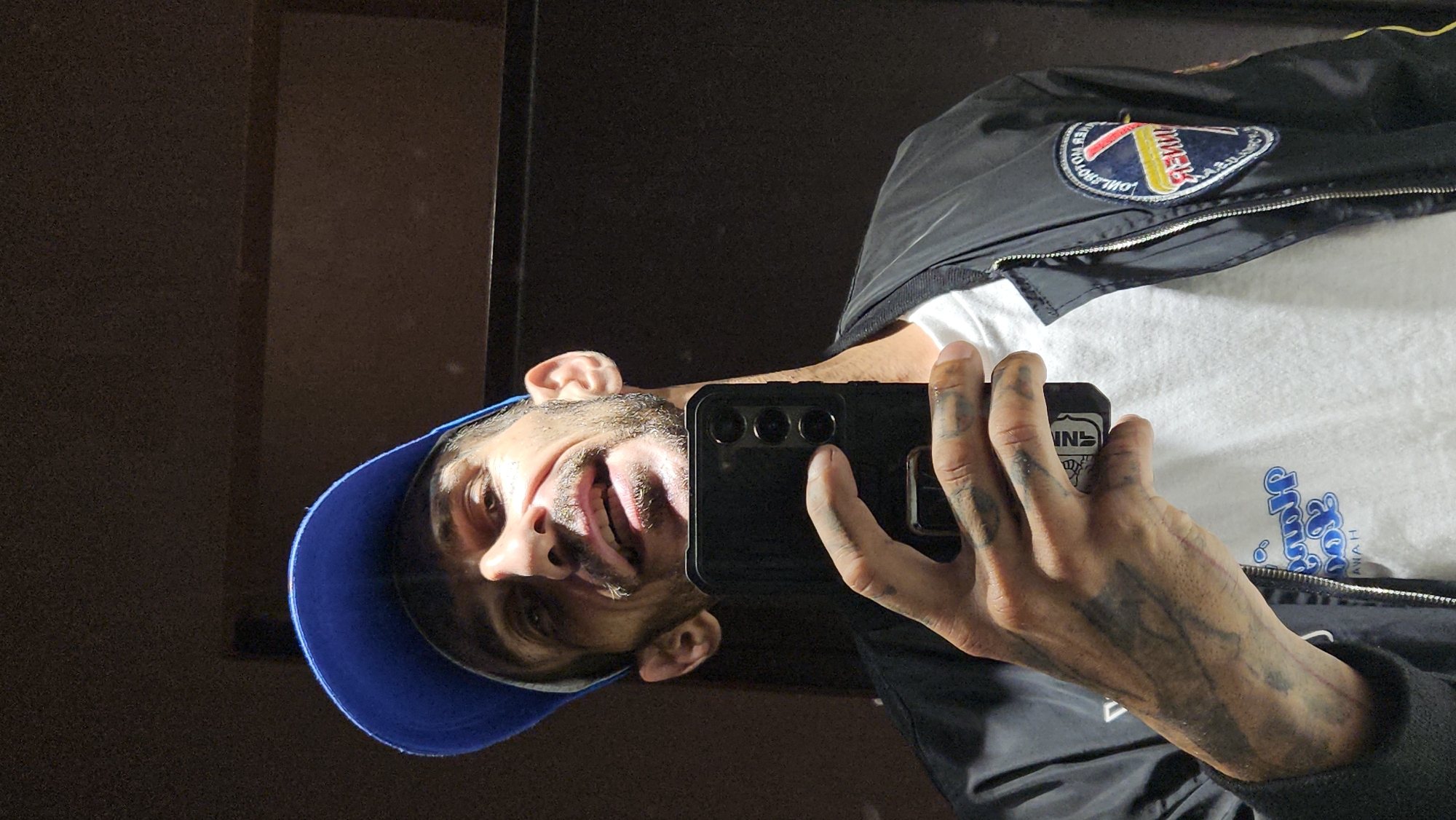 A man with light brown skin, a beard, and a blue baseball cap is taking a selfie in a mirror. He's holding a black smartphone with a triple-lens camera setup. His left hand is visible and covered in tattoos. He is wearing a white t-shirt and a black bomber jacket with a circular patch on the right shoulder. The reflection shows a dark wall behind him. The photo gives no indication of the city of Nha Trang, Vietnam.