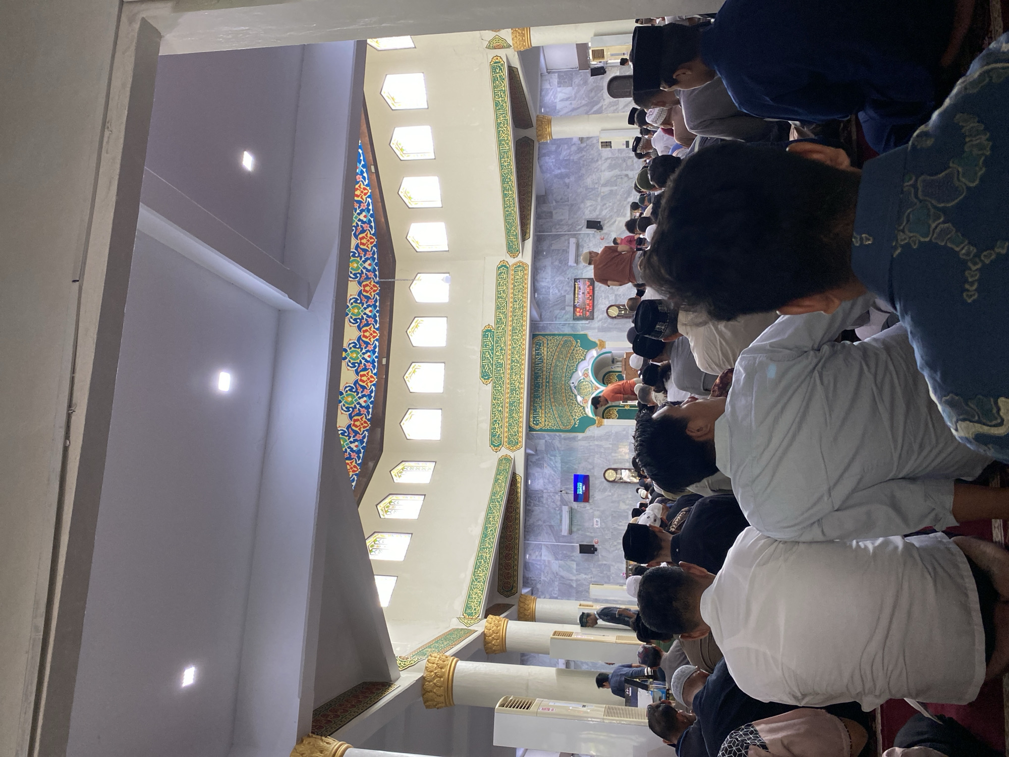 The image depicts a gathering of people inside a mosque, likely during a prayer service. The perspective is from a high angle, looking down into the prayer hall. Many individuals are visible, dressed in traditional attire, including kufis and modest clothing. They appear to be in various prayer postures, suggesting an ongoing religious observance.
The interior of the mosque is ornately decorated. The walls feature intricate Islamic calligraphy in green and gold, interspersed with colorful patterned friezes. Large, decorative pillars with gold accents support the ceiling. Natural light filters in through a series of rectangular windows arranged along the upper walls. The floor is covered with prayer mats. In the background, a television screen displays an image, and an air conditioning unit is mounted on the wall.
The scene is well-lit, suggesting it is daytime. The overall atmosphere appears serene and devout. While no specific time of day is explicitly indicated, the presence of multiple people engaged in prayer points to a communal religious activity. The location context, Lhokseumawe, Indonesia, implies a Muslim place of worship. No discernible weather conditions are evident from the indoor setting.