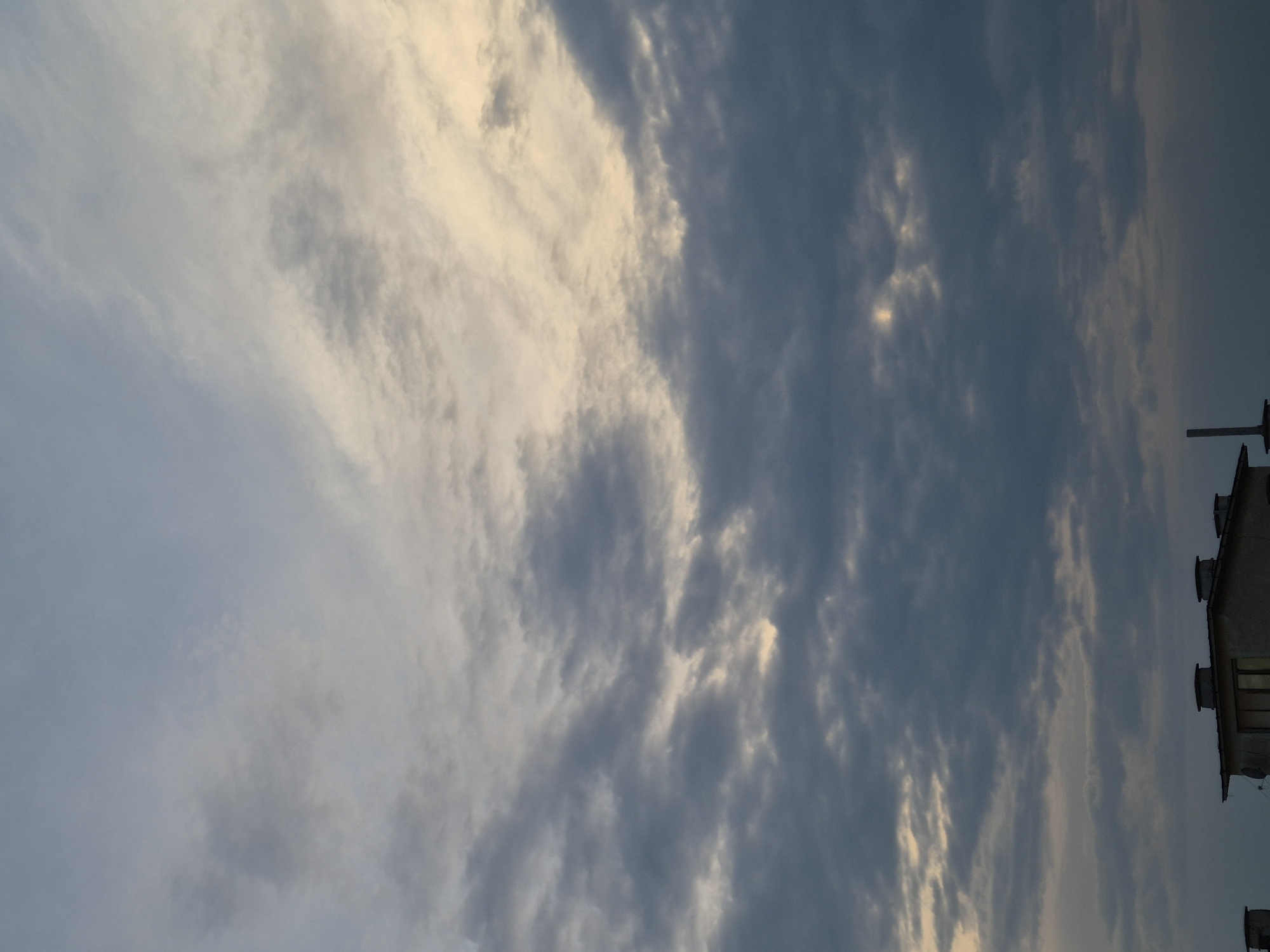The image displays a sky scene dominated by an expanse of clouds, with a partial view of a building visible along the right edge. The sky transitions between various shades of blue-grey and areas of bright, warm golden-yellow light, indicating either sunrise or sunset. Wispy, translucent clouds intermingle with more dense and textured formations.
On the right side of the frame, the roof and a small portion of the upper section of a building are visible. Several rectangular structures, possibly vents or air conditioning units, are distributed across the rooftop. A distinct, dark, vertical pole or antenna also extends upwards from the roof. The building itself appears to be light-colored with a dark roof. No people are visible, and no specific activity is taking place. The weather conditions suggest a partly cloudy sky with the sun low on the horizon. There is no visible text in the image.