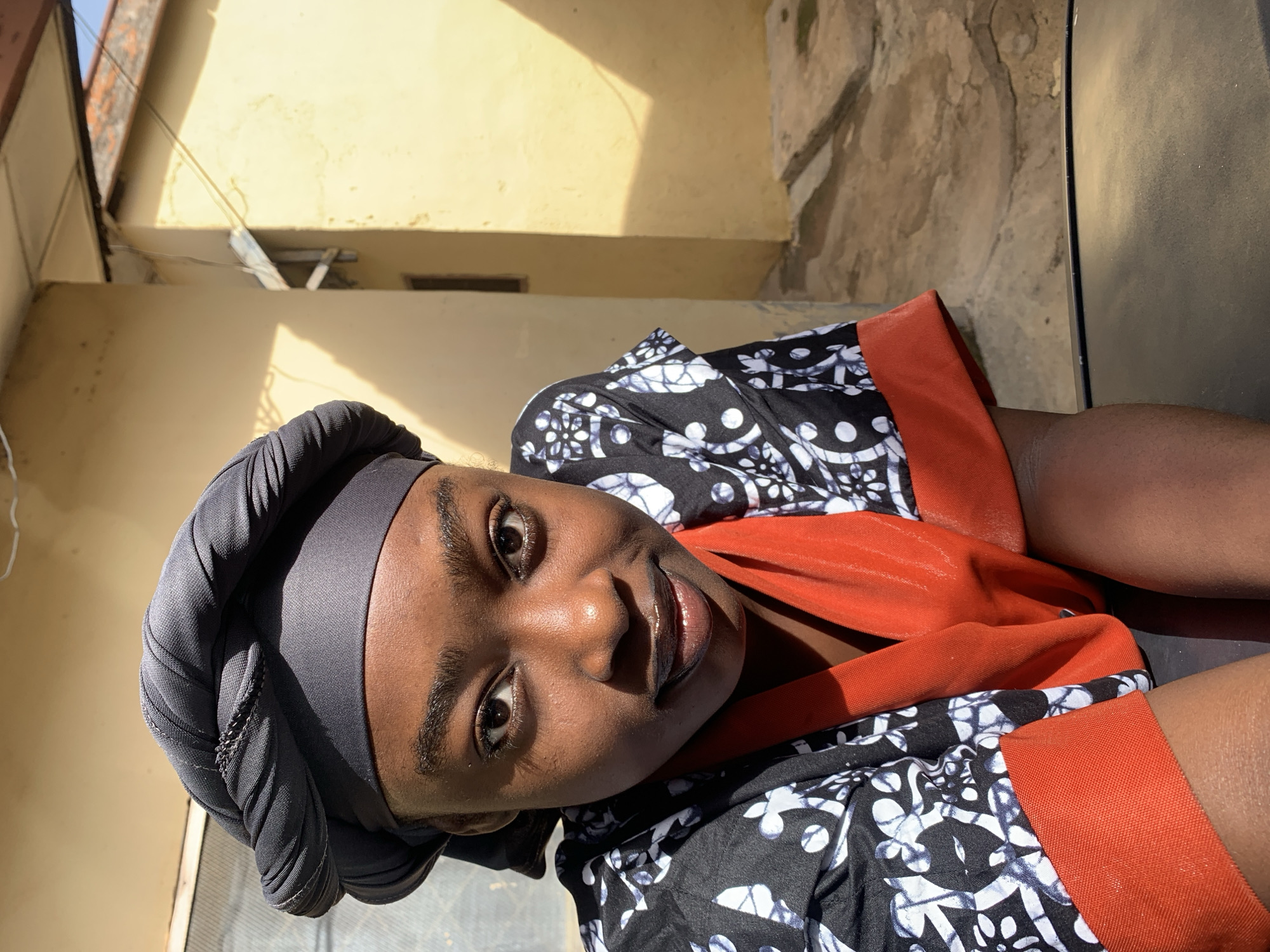 A young woman with dark skin and a black headscarf is pictured in Rafin Par, Nigeria. She is wearing a black and white patterned garment with orange trim, and her face is turned towards the camera. Sunlight illuminates the right side of her face and the yellow building behind her. The setting appears to be an outdoor area with concrete ground and a portion of a building visible.