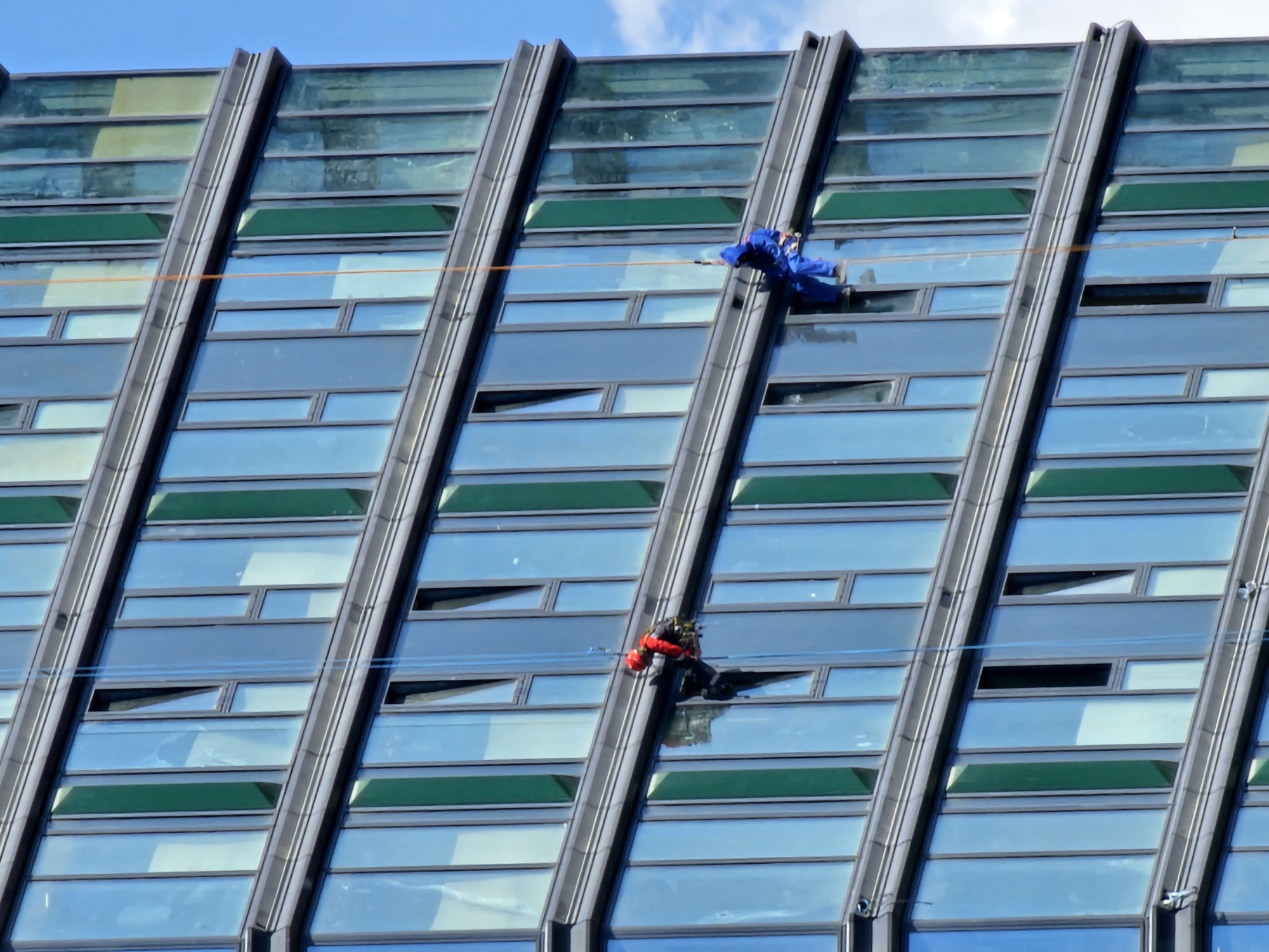 The image shows two window washers rappelling down the side of a modern skyscraper. The building has a glass facade with alternating sections of clear blue glass and dark green panels. The window washers are suspended from ropes attached to the top of the building. One washer, wearing a red jacket and helmet, is on a lower level, appearing to be working. The other washer, wearing a blue jumpsuit, is on a higher level, also appearing to be working. Both are attached to safety harnesses. The sky is blue with a few white clouds visible. The scene depicts a high-rise maintenance operation in an urban environment. The architectural style of the building suggests a contemporary urban setting, but the location in Moscow, Russia cannot be determined from the image alone.